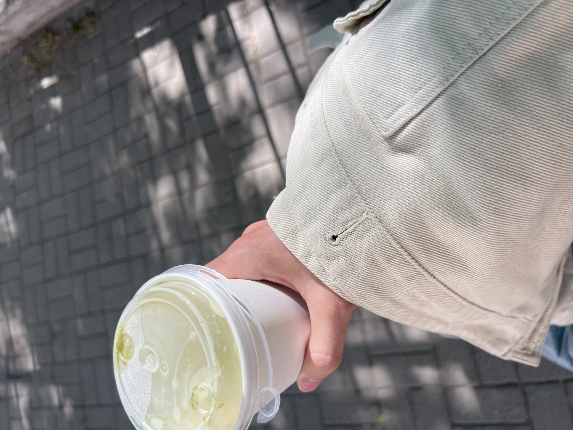 A person is holding a disposable cup with a clear plastic lid containing a yellow liquid, likely a beverage. The person's hand is around the cup, and they are wearing a light-colored jacket. The background shows a gray brick sidewalk with shadows cast by trees or other objects. The scene appears to be outdoors, possibly in an urban area. The location is not discernible from the image.