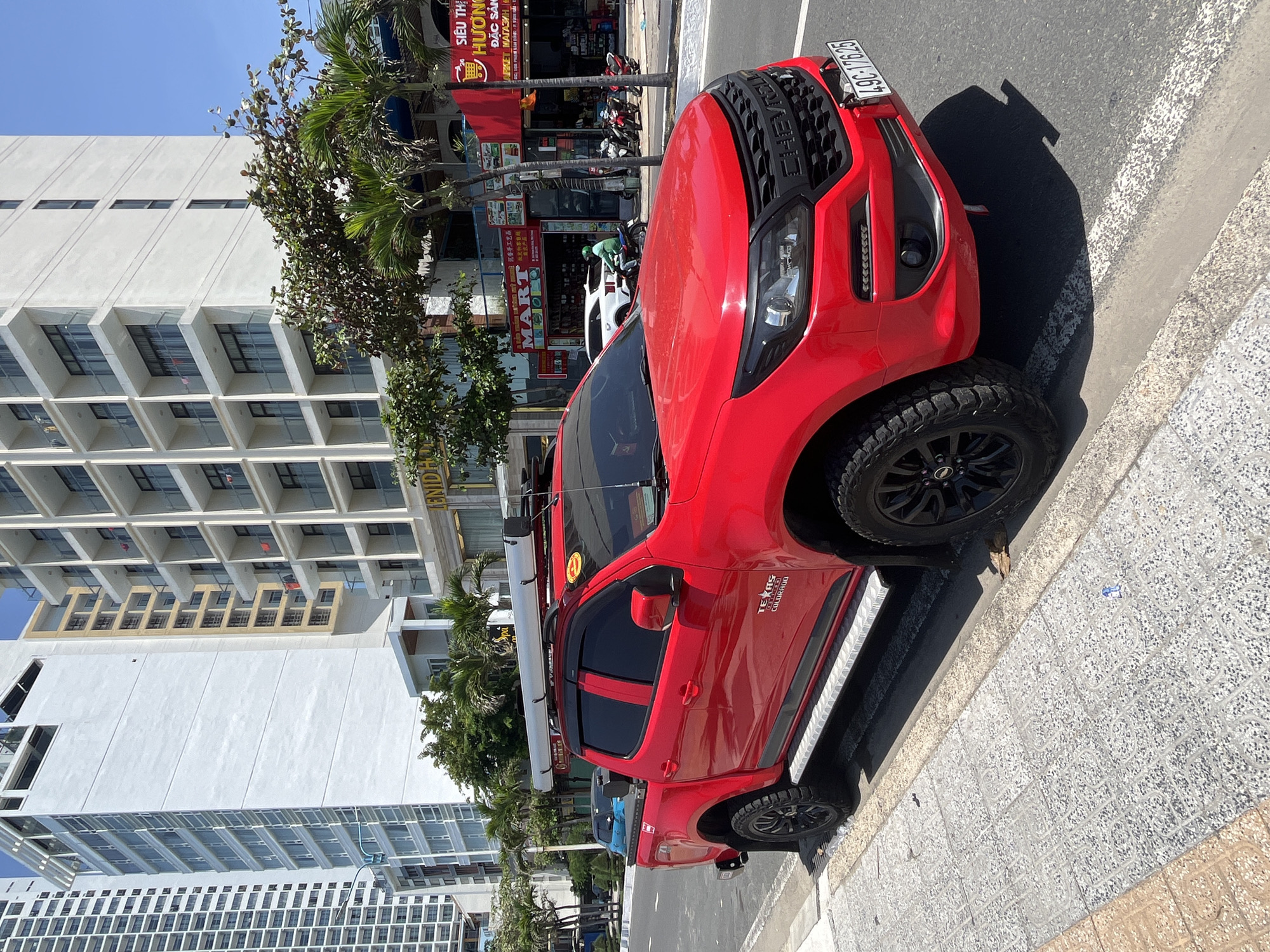 The image depicts a red Chevrolet Colorado pickup truck parked on the right side of a sunlit street, partially on the sidewalk, in Bac Nha Trang Ward, Vietnam. The truck is customized with a lift kit, black multi-spoke wheels fitted with aggressive off-road tires, diamond plate side steps, and dark tinted windows. A roof rack supports a long, silver cylindrical object, possibly an awning or storage tube, above the cab, and a blue container is visible in the truck bed. The vehicle bears a Vietnamese license plate, "49C-176.25," attached to the front bumper.
In the background, a bustling urban scene unfolds. A tall, modern white high-rise building with numerous windows dominates the left side. To the right, several multi-story buildings with balconies line the street, one prominently displaying "LENDI HOTEL" on its ground-level signage. Further right, storefronts are visible, including one marked "MART" and another with Vietnamese text indicating a "S siêu thị HƯƠNG" (HƯƠNG Supermarket), both featuring red and white signage. Palm trees and other tropical foliage line the sidewalk, casting shadows. On the road, a white SUV is parked in front of the shops, and a person on a green scooter is riding past. Several motorbikes are parked near the storefronts. The sky is clear blue, indicating bright, sunny weather. The sidewalk is paved with patterned grey tiles, with a small blue wrapper visible on its surface.