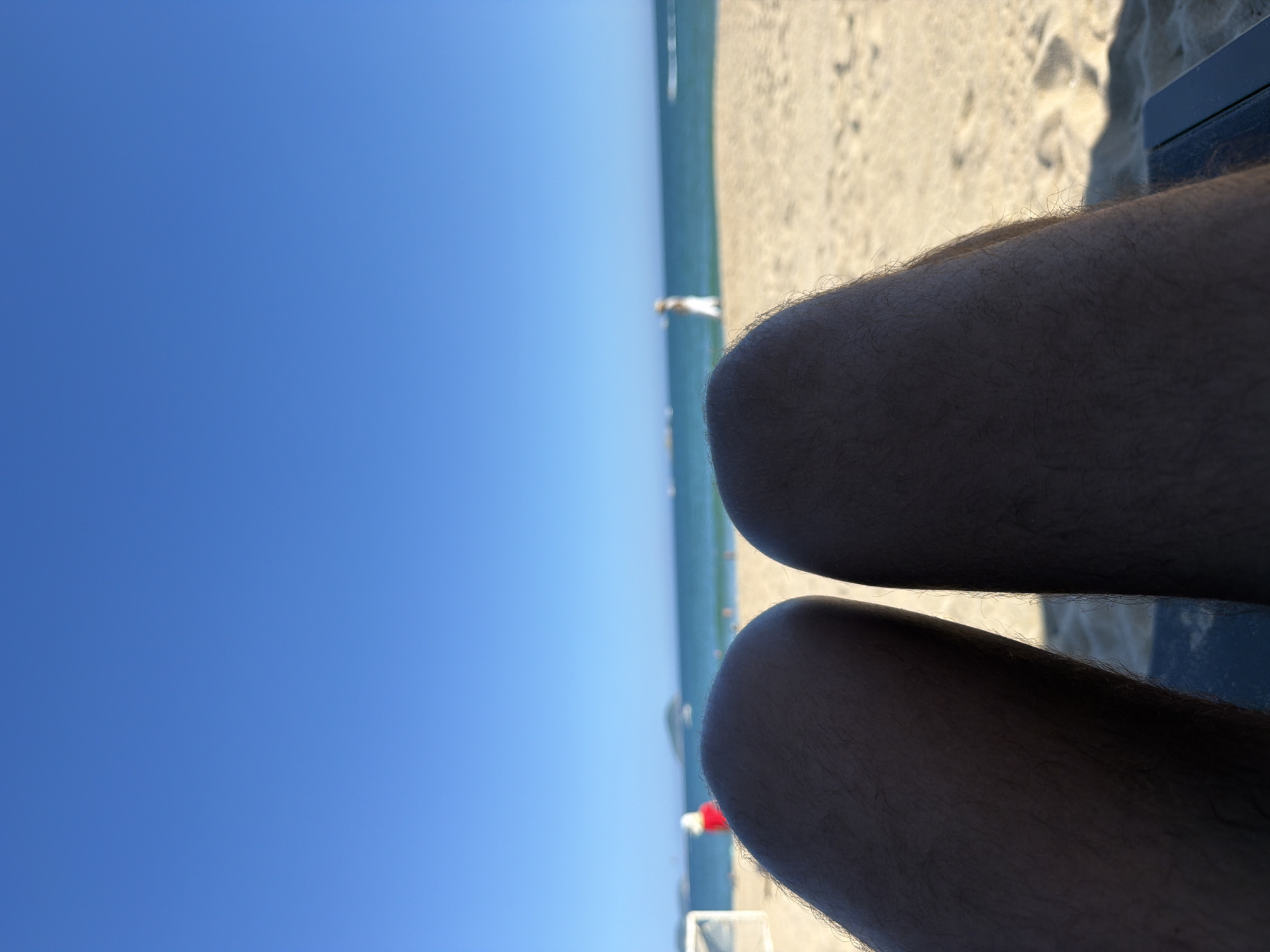 A first-person perspective shot shows the legs of a person sitting on a beach in Jiyang District, China. The legs are close to the camera, filling the lower half of the frame. Beyond the legs, the scene opens up to a sandy beach with a clear blue sky above. The ocean stretches out to the horizon, with a few distant islands or landmasses visible. In the middle ground, a person in a white hat and red clothing stands on the beach, and another person in a white dress is further away near the water. The sun appears to be bright, casting shadows on the sand.