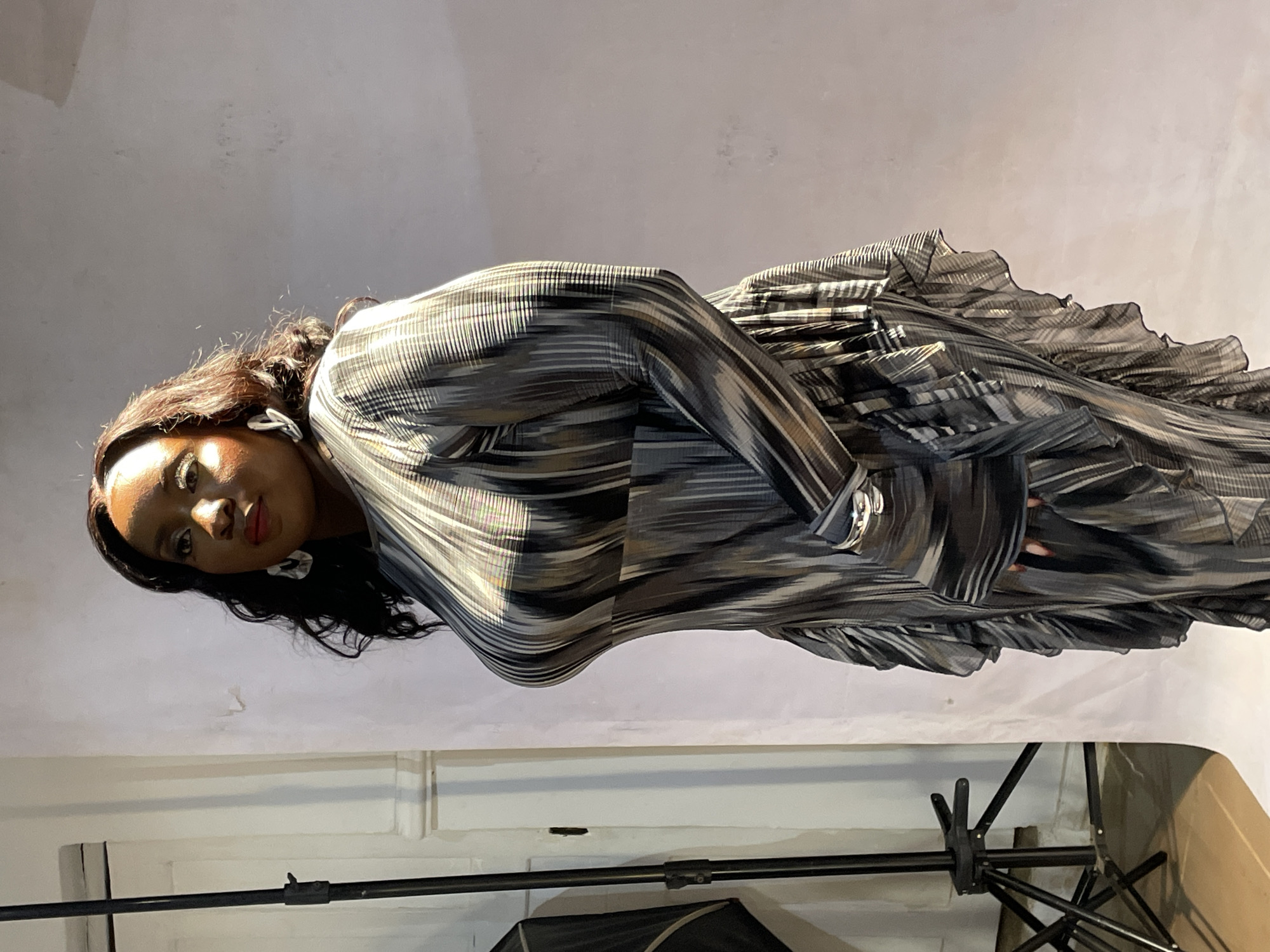 A woman with dark skin and long, dark hair is captured in a full-body shot, posing for a photograph. She looks slightly off-camera with a composed expression, wearing red lipstick and large, light-colored earrings. She is dressed in a long-sleeved, full-length dress featuring an intricate pattern of horizontal and diagonal stripes in shades of grey, black, brown, and white. The dress appears to have a pleated or ruffled skirt, adding texture and volume.
The setting is indoors, with a plain, light grey or off-white wall serving as the background. The lighting is artificial, creating highlights on the woman's shoulder and the textured fabric of her dress. In the lower right portion of the image, black studio equipment, including a tripod-like stand and a dark softbox or umbrella, is partially visible, indicating a professional photoshoot environment. No specific time of day or weather can be determined from the indoor setting and artificial lighting. There is no visible text in the image. The event appears to be a fashion or portrait photoshoot taking place in Barakin Akawo, Nigeria.