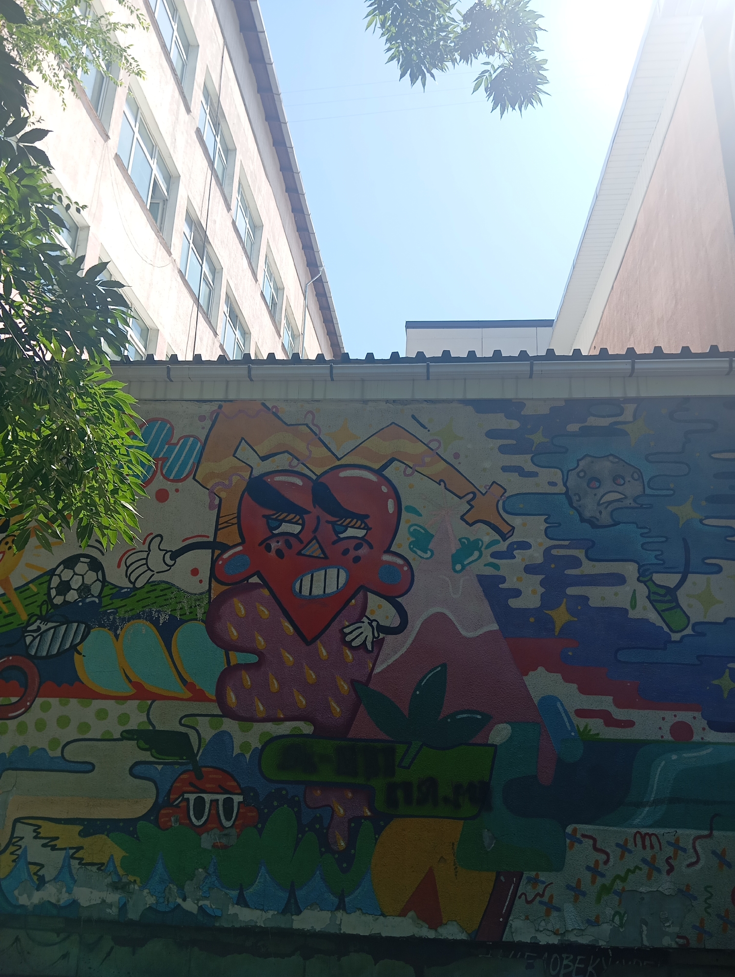 The image shows a colorful mural on a wall in Almaty, Kazakhstan. The mural features cartoonish characters, including a distressed red heart with large eyes and a grumpy moon-like face. There are also abstract shapes, stars, and a soccer ball. The wall appears to be located in an urban setting, possibly an alleyway or the side of a building, with trees and a bright blue sky visible above. The lower part of the wall shows signs of wear and tear, with peeling paint. The overall impression is one of vibrant street art contrasting with a somewhat aged urban environment.