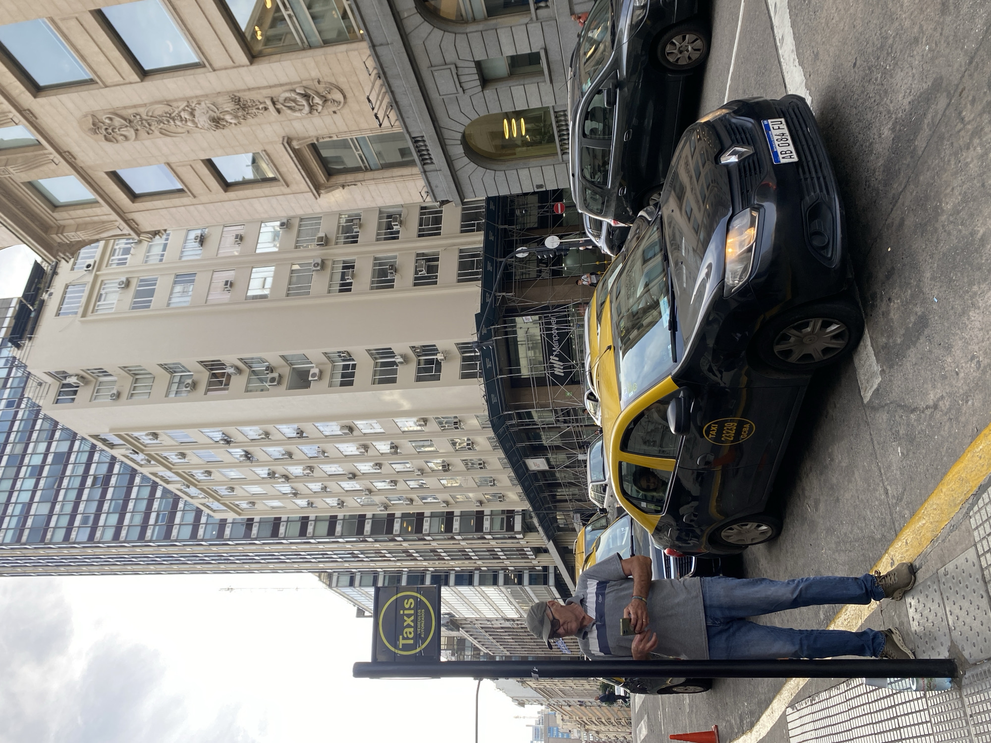 The image captures a street scene in Buenos Aires, Argentina. A man stands on the sidewalk near a black "Taxis Autorizados" sign, holding a smartphone. Yellow and black taxis are lined up along the curb. Behind the taxis, scaffolding covers part of a building under construction or renovation. Buildings with classical architectural details and numerous windows line the street. The license plate on the front taxi is visible and reads "AB 084 EU". The environment suggests an urban setting with a mix of old and modern architecture. The sky is overcast.
