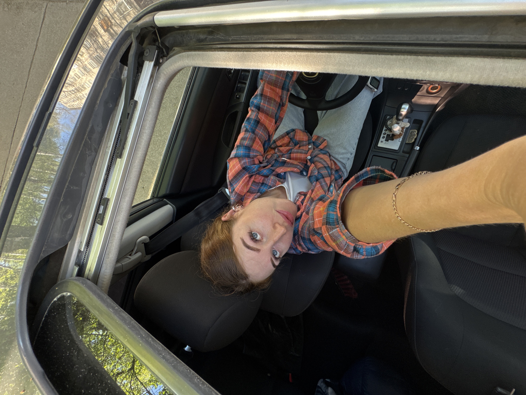 A woman is sitting in the driver's seat of a car with an open sunroof, taking a selfie. She is wearing a plaid shirt and light-colored pants. The interior of the car is visible, including the steering wheel, dashboard, and seats. The location is not possible to verify in the city of Chaikovsky, Russia from the current image. The environment outside the sunroof shows trees and possible buildings.