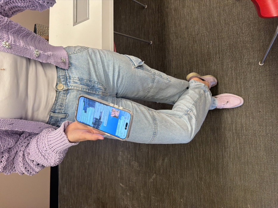 A person sits with crossed legs wearing light blue jeans, a white top, and a lavender cardigan. They hold a smartphone displaying a chat screen. The scene is lit by artificial light, casting a neutral tone. The background features a dark grey carpet with a linear pattern and glimpses of furniture, including the edge of a white desk and a red chair. Lavender shoes are visible on the person's feet.