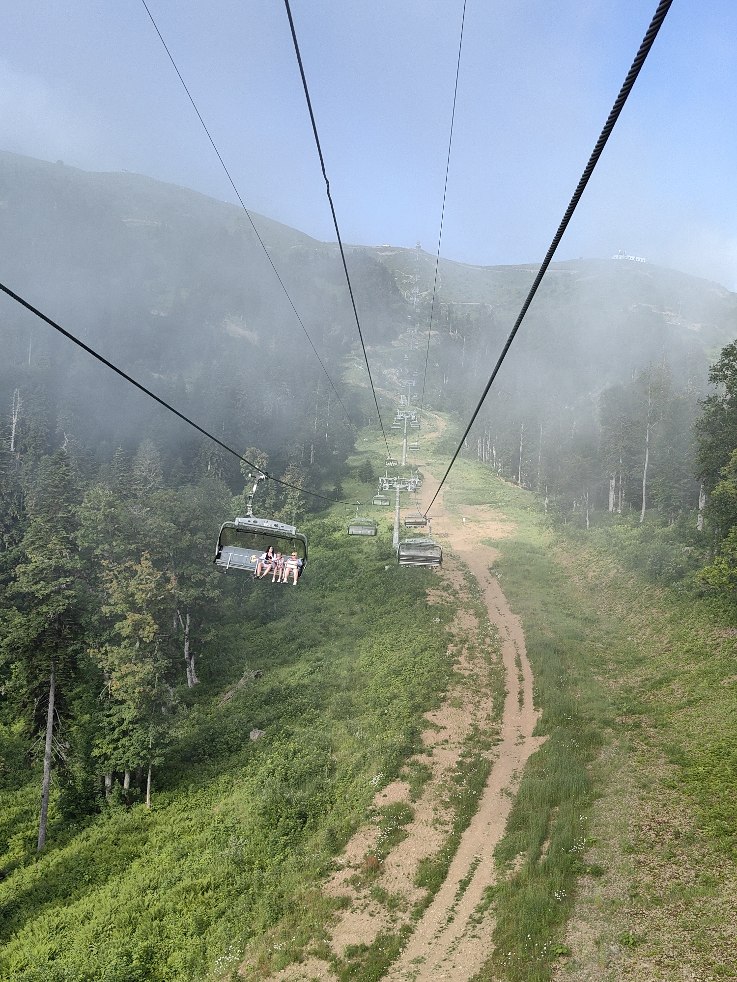 This image depicts a view from a ski lift in Estosadok, Russia. The ski lift is ascending a mountainside covered in lush green vegetation and trees. In the foreground, a chair on the ski lift carries four individuals. Further up the mountain, more ski lift chairs are visible, some empty and some with passengers. A dirt path winds its way up the slope next to the ski lift. The sky is a clear blue with some wisps of fog or clouds obscuring the distant mountain peaks. The scene suggests a summer or early autumn day, as there is no snow on the ground.