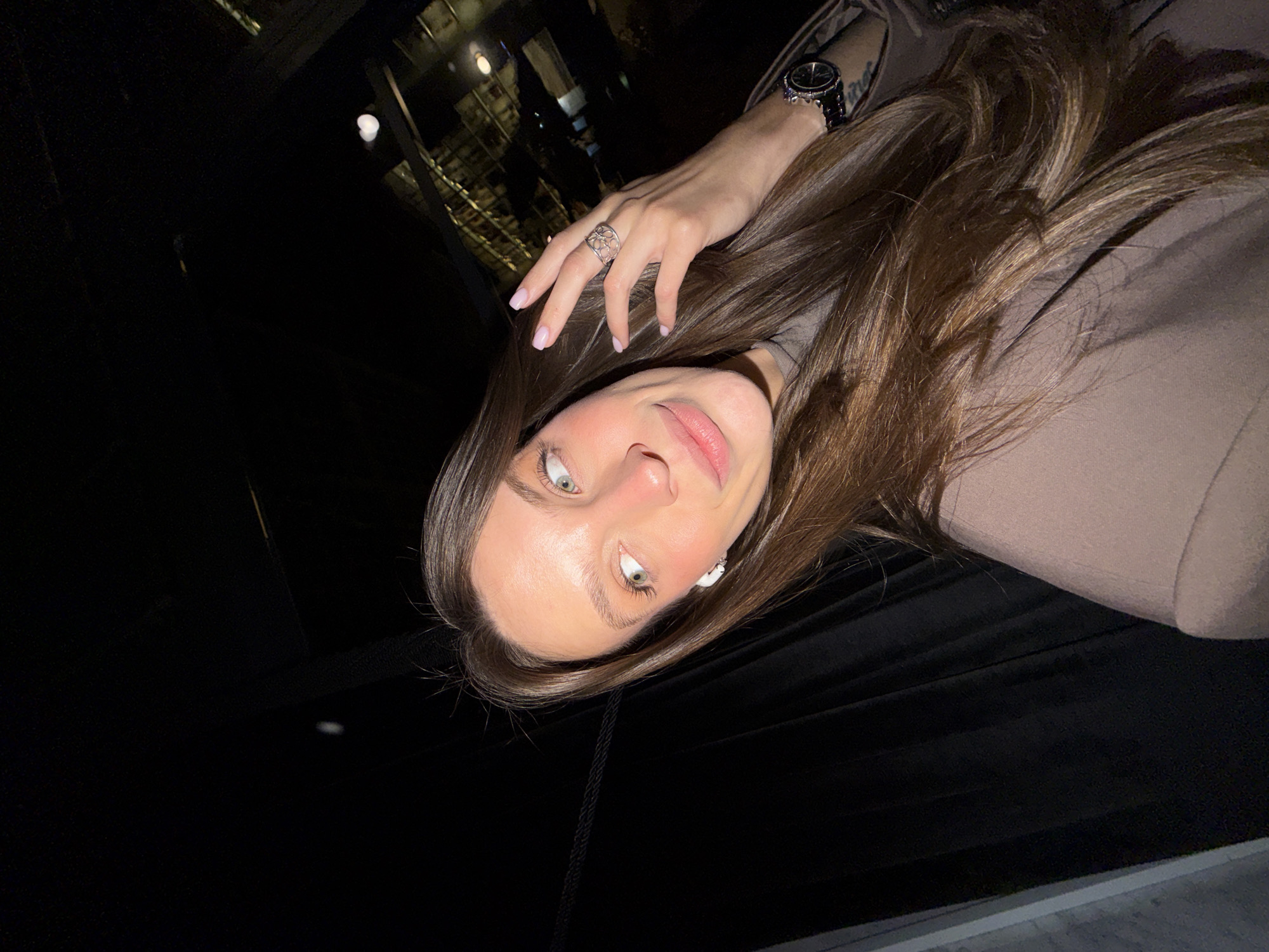 A selfie of a woman with light skin and green eyes, facing the camera at an upward angle. She has long brown hair. The woman wears a beige jacket. She has a ring on one hand and a watch on the other. The background is dark, but suggests a restaurant setting with a staircase in the upper left. The precise location in Budva, Montenegro, is not determinable from the provided image.