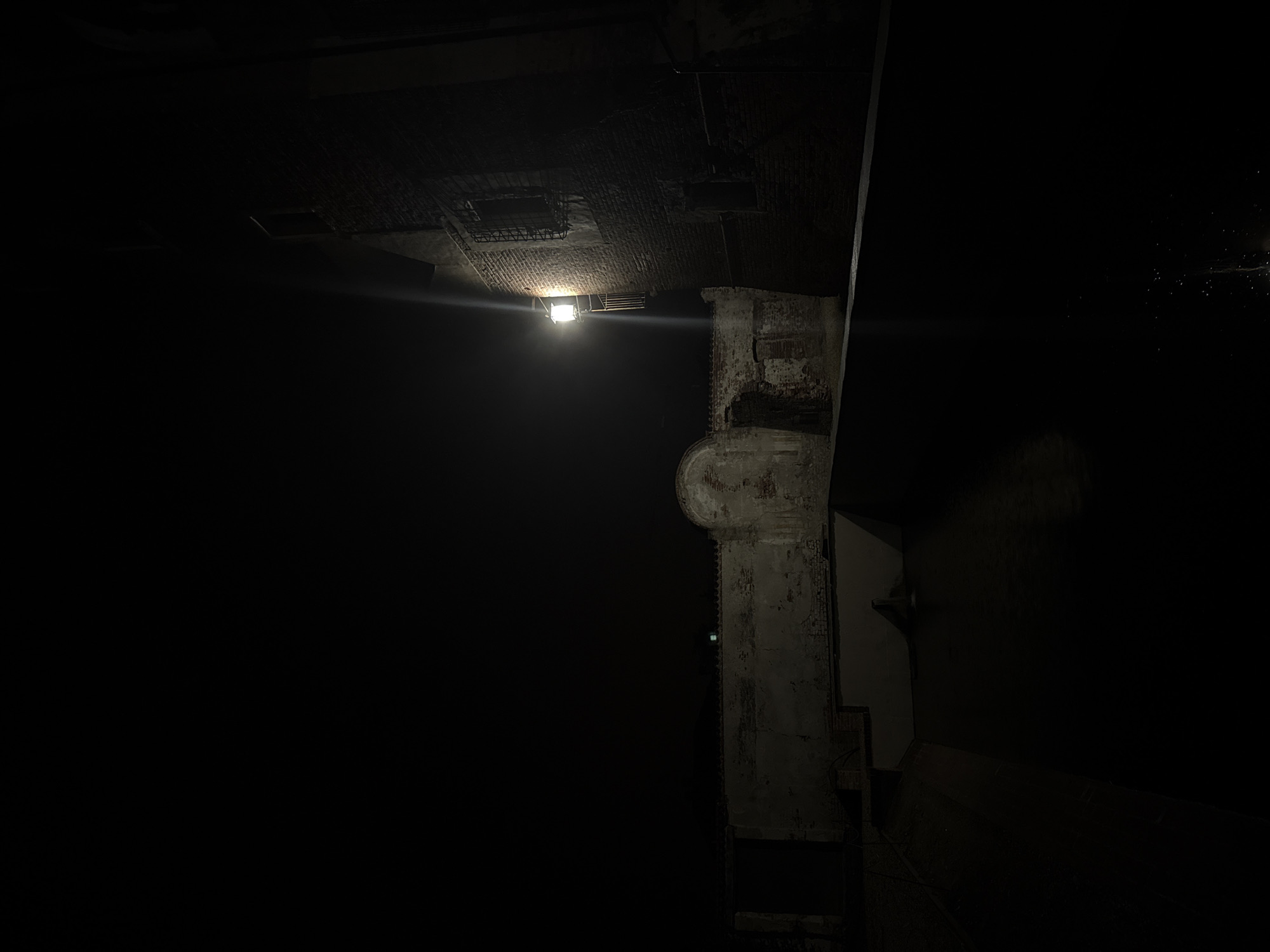 The image depicts a dimly lit, exterior scene at night, likely in the historical center of Lagnasco, Italy. The foreground is dominated by a dark body of water, possibly a canal or fountain, with small points of light reflecting on its surface.
To the left, a weathered, aged wall runs horizontally across the frame. This wall features architectural elements like a partially visible arched entryway and sections where the brickwork is exposed. The wall appears to be part of an older structure, possibly a courtyard or a protected area.
To the right, a brick building stands tall, its facade illuminated by a single, vintage-style lantern mounted on the wall. This lantern casts a bright, focused beam of light that creates a distinct streak of illumination against the dark night sky. The building itself has a window with a barred grate, suggesting historical security measures or architectural design. The brickwork appears rough and aged, consistent with older construction.
The overall atmosphere is one of quiet solitude and historical ambiance, enhanced by the nighttime lighting and the aged structures. The scene suggests a place steeped in history, where the passage of time is evident in the materials and architecture. There are no people visible in the image.