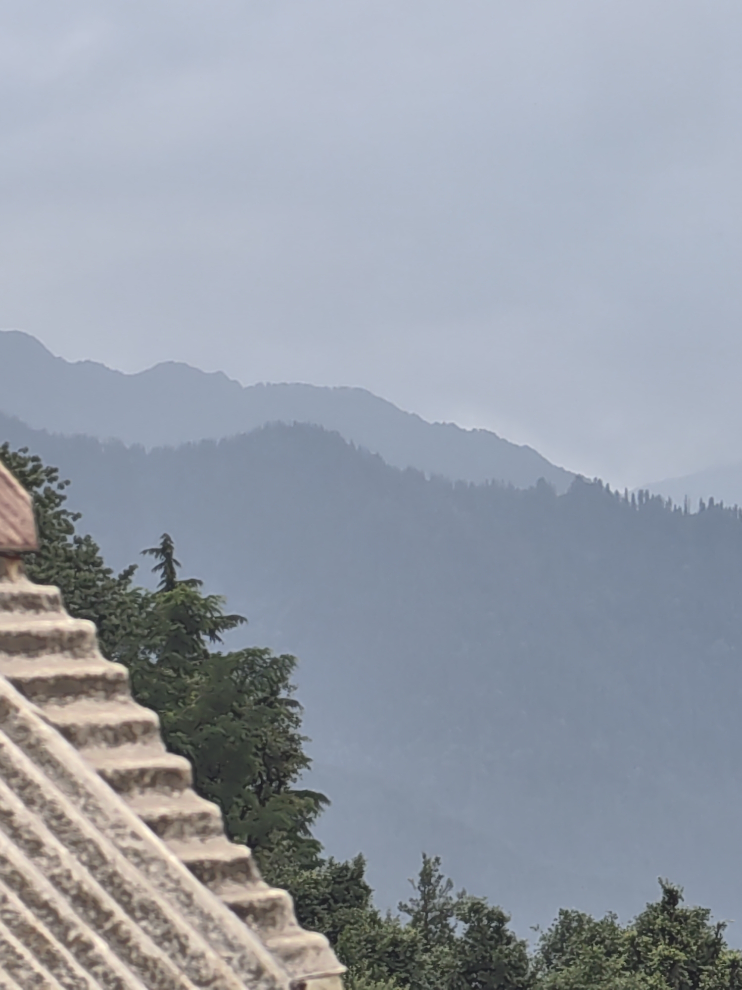The image displays a layered view of mountainous terrain, characteristic of Vashisht, India. In the foreground, a textured, stepped structure, possibly a rooftop or terrace, occupies the lower left quadrant. Beyond this structure, a dense collection of green trees forms a verdant band across the mid-ground. The background is dominated by the hazy silhouettes of multiple mountain ranges, fading into a muted sky. The overall atmosphere is serene and suggests a natural, possibly elevated landscape.