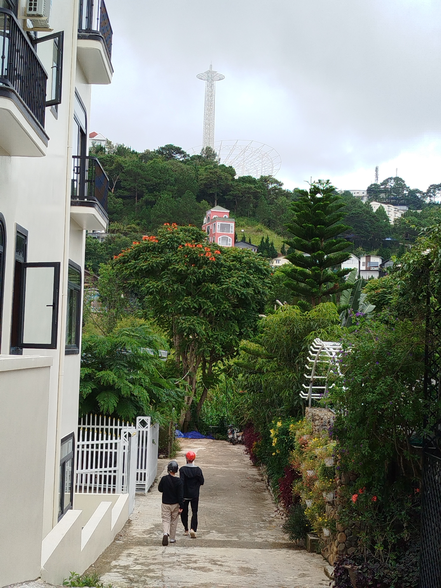 The image depicts a view looking down a paved pathway in Da Lat, Vietnam. On the left, a white building with black trim and balconies is prominently featured. Two individuals are walking away from the camera down the pathway, one wearing a red helmet and the other a black helmet. The pathway is bordered by a white picket fence and lush green foliage. In the background, a distinctive pink building with white trim is visible amidst a hillside covered in trees. Further in the distance, a tall, slender white tower structure, possibly a radio tower or observation tower, rises above the treeline. The sky is overcast, suggesting a cloudy day. The overall scene conveys a sense of a residential area or tourist destination in a hilly, green environment.
