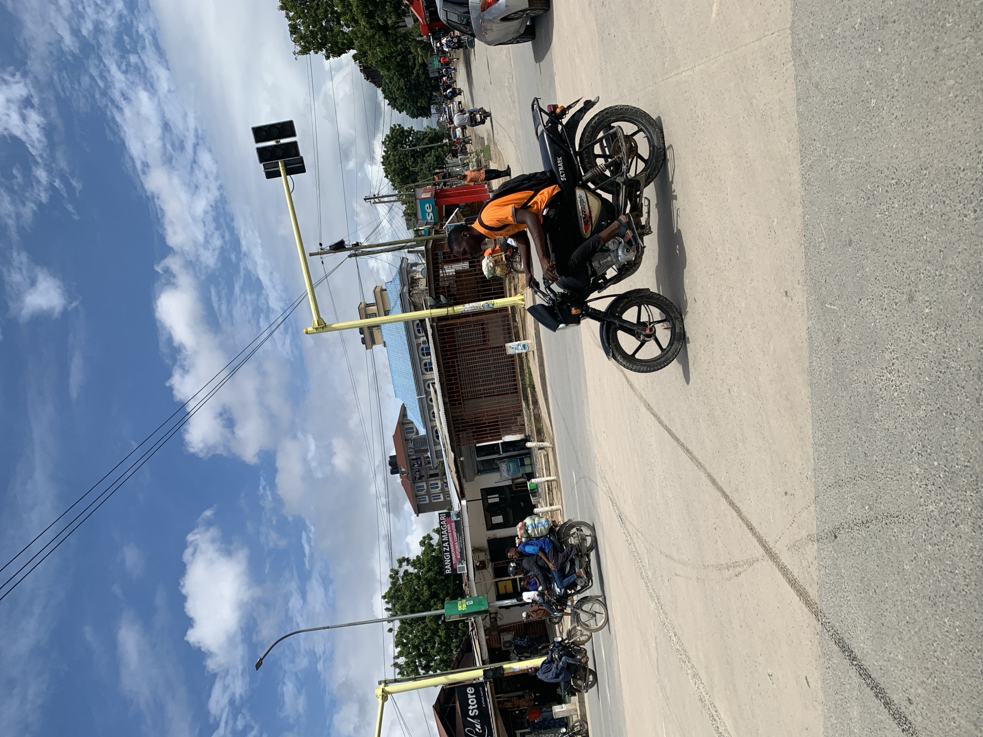 A man in an orange shirt is seated on a black SkyMark motorcycle at an intersection in Dar es-Salaam, Tanzania. There are tire marks on the pavement. In the background, traffic waits under a traffic light. Businesses are visible, including a shop front with "RANGI ZA MAGARI" written on it. The sky is partly cloudy.