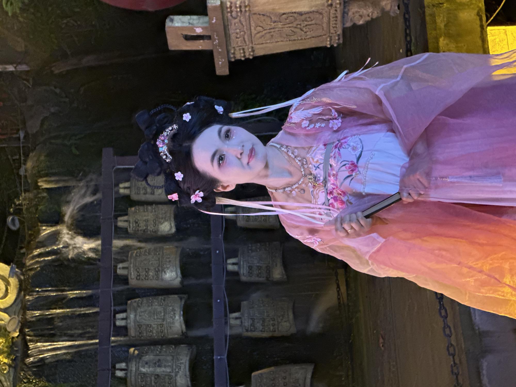 A low-angle, medium shot shows a person wearing a pink traditional Chinese hanfu, standing near a waterfall in Yuzhong District, Chongqing, China. The person has black hair styled in an updo with floral hair ornaments and is wearing a pearl necklace and makeup. They are holding a fan in their hands. Behind the person, a waterfall flows down a wall with metal objects attached. To the left of the person, there is a dark background. The image is well-lit, with the person and the waterfall being the main focus.