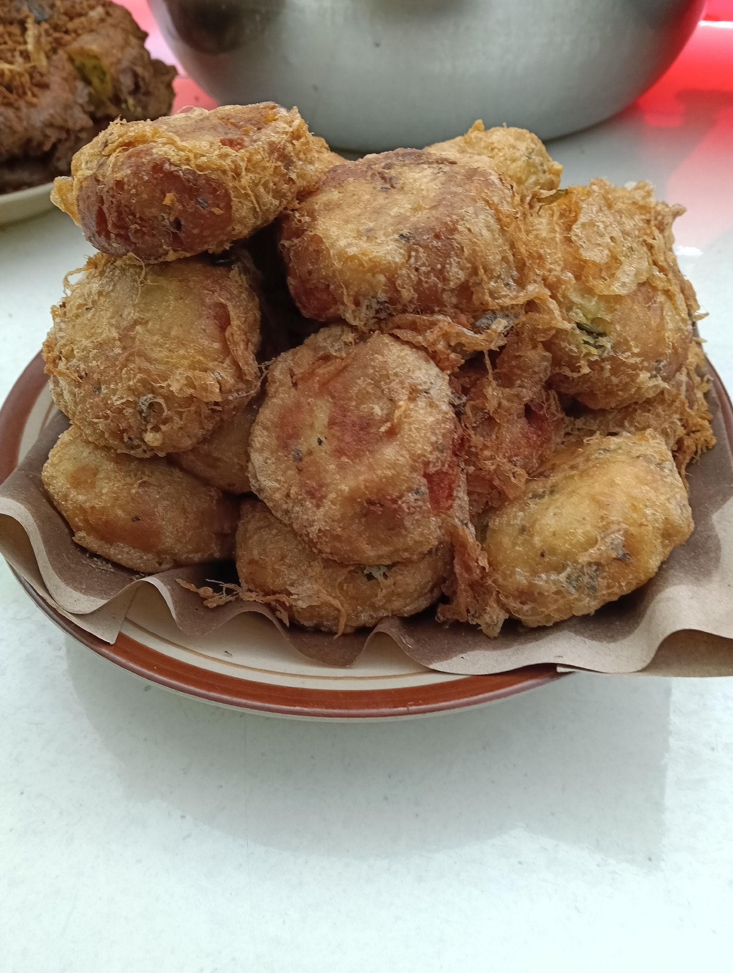 A plate of fried food, likely fritters or patties, is presented on a light-colored textured surface. The food items are golden brown and appear to be somewhat irregular in shape, piled together on the plate. The plate itself has a brown and white striped rim. Underneath the food, brown paper is visible, adding to the rustic presentation. In the background, the edge of a large metal bowl or pot is partially visible, along with a blurry pinkish-red element and a dark brown food item with shredded toppings. The lighting suggests an indoor setting. The scene is likely a food presentation in Banjarnegoro, Indonesia.