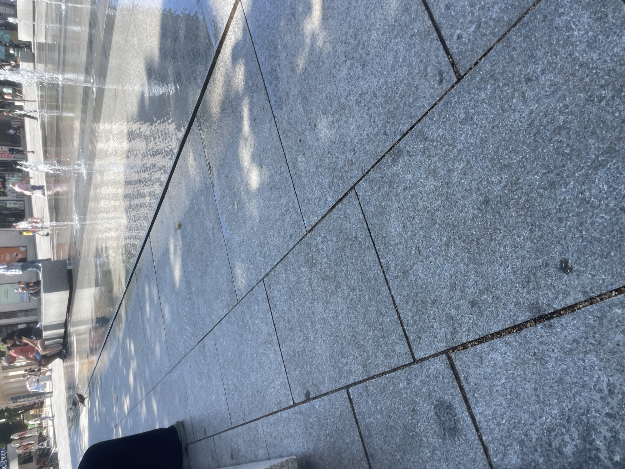 The image depicts a view of a pedestrian area in the city of Stara Zagora, Bulgaria, featuring a modern fountain. In the foreground, large granite tiles are laid out, showing subtle water reflections and shadows from surrounding trees and structures. To the left, a person's dark-clad legs and light-colored shoes are visible, suggesting they are sitting or resting.
In the mid-ground, a shallow fountain area is active, with jets of water creating splashes and pools. Several pigeons are present in the fountain area, with one actively walking on the wet pavement. Beyond the fountain, a group of people are seated on a low, wide concrete structure, likely a bench or part of the fountain's design. One individual wears a red shirt and blue shorts, another a black t-shirt and skirt.
In the background, the scene transitions to a streetscape with buildings, shops, and more pedestrians. The architecture appears to be a mix of modern and older styles. The overall atmosphere suggests a bright, sunny day in a public urban space. The image composition places a strong emphasis on the textured pavement and the interplay of light and water.