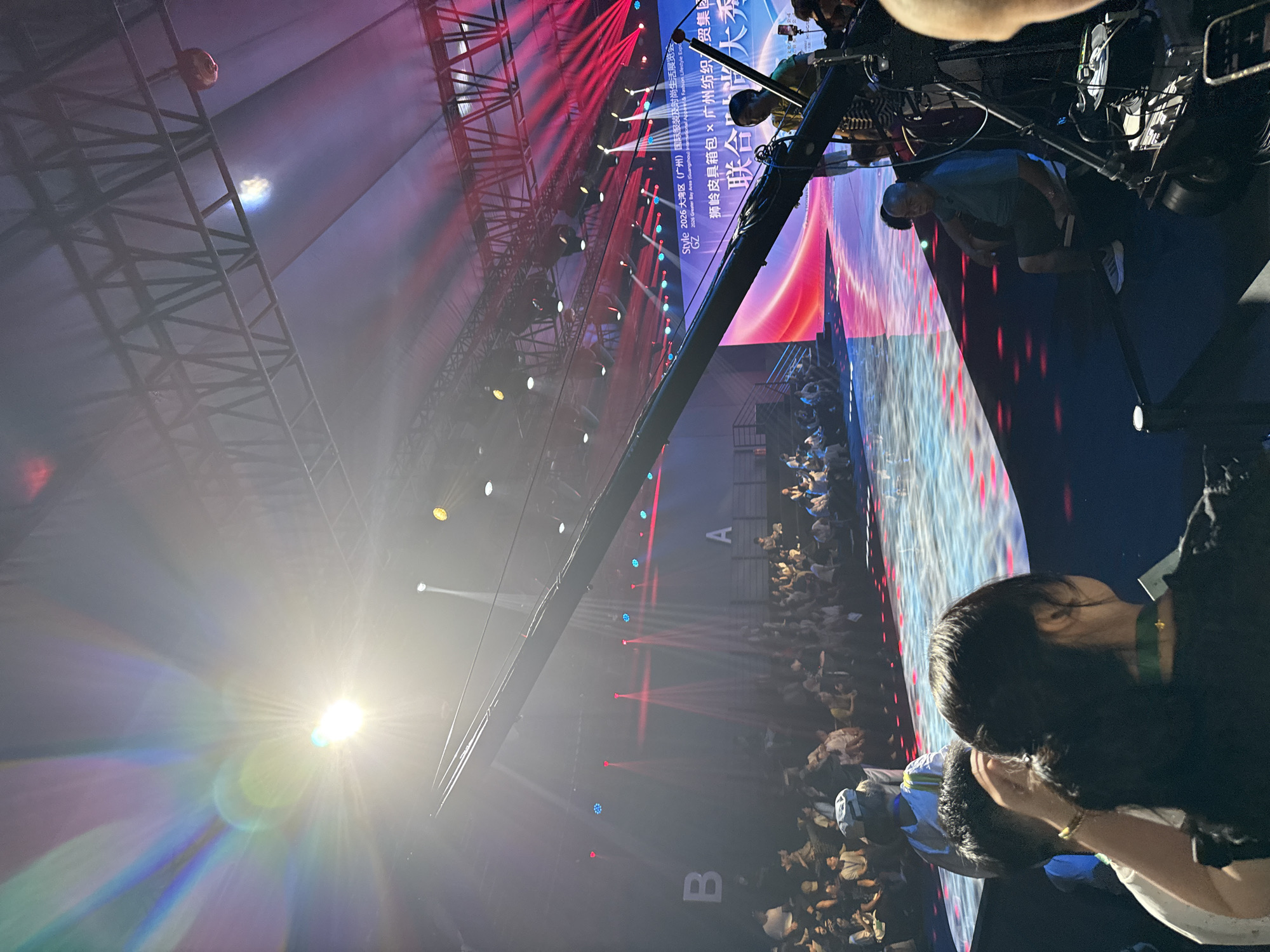 A wide-angle, eye-level outdoor shot captures an indoor event with a brightly lit stage and audience seating. The scene appears to be a fashion show or similar exhibition, evidenced by the runway, seated audience, and stage lighting. The foreground is dominated by a large camera jib, with its long arm extending towards the stage, and a camera operator sitting on a stool, positioned to the right of the frame. In the background, a large digital screen displays event information, including "STYLE 2026 大湾区 (广州) 国际服装时尚生活展会 GZ 2026 Greater Bay Area (Guangzhou) International Apparel & Fashion Lifestyle Expo" and Chinese characters. The audience is seated in tiered rows on both sides of the stage area. The stage itself is illuminated with dynamic colored lights, primarily red and blue, with beams of light creating dramatic effects. The overall atmosphere suggests a professional production, likely taking place in a convention center or exhibition hall within Haizhu District, China.