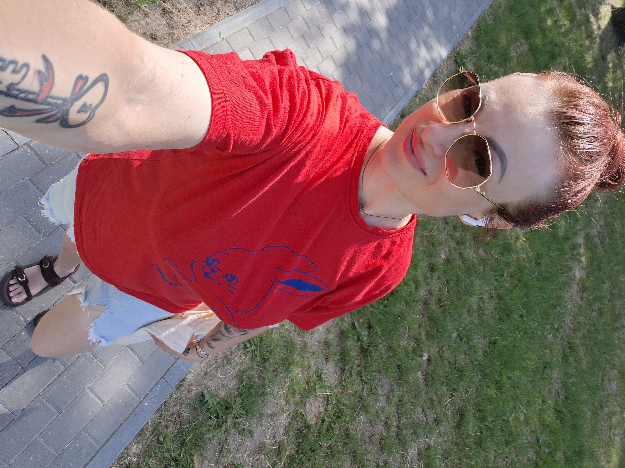 A woman is taking a selfie outdoors on a sunny day. She has red hair pulled up in a bun, wears sunglasses, and has wireless earbuds in her ears. She is dressed in a red t-shirt with a blue bunny graphic and light-wash denim shorts. She is wearing dark brown sandals. A canvas bag is visible. Her left arm has tattoos. She is standing on a paved walkway next to a grassy area. The location is identified as Забалоцкі сельскі Савет, Belarus.