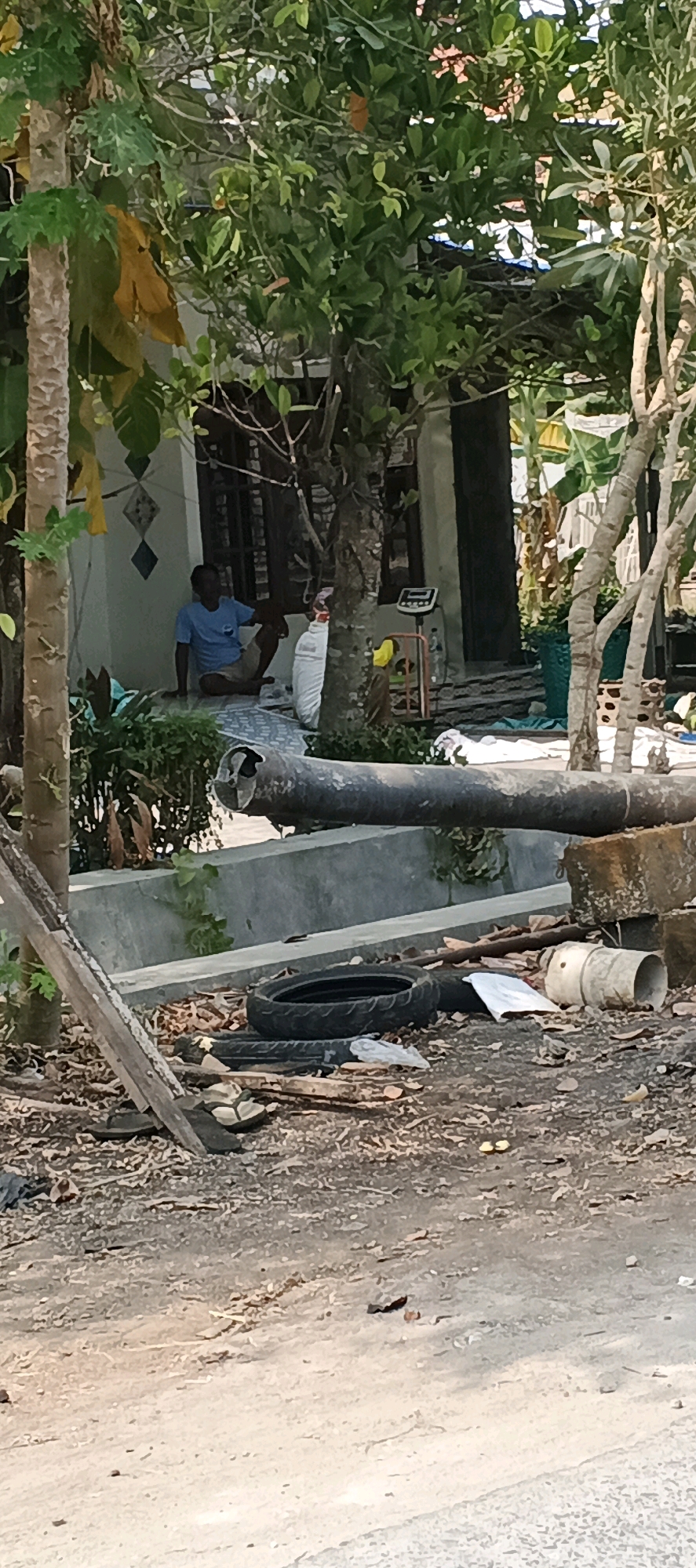 In Latukan, Indonesia, a man in a blue t-shirt and tan shorts sits on a patterned mat outside a house with white walls and dark wooden window frames. He is looking towards the left side of the frame. In front of the house, a large, broken concrete pipe lies horizontally across the scene. The ground is covered in dry leaves, debris, and some scattered tires. To the right of the pipe, a white cylindrical object and what appears to be a white plastic bag are visible. The environment is outdoors, with abundant greenery from trees and foliage surrounding the area. The overall scene suggests a somewhat disheveled outdoor space.
