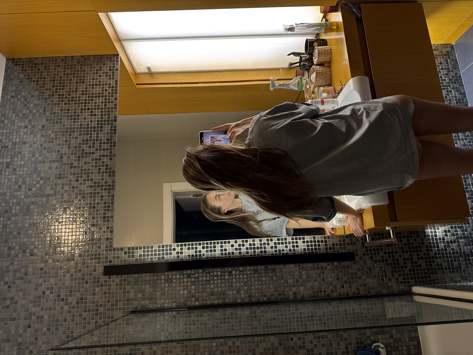 A woman with long, light-brown hair is taking a photo of her reflection in a mirror in a bathroom. She is holding a smartphone horizontally. She is wearing a gray oversized t-shirt. The bathroom has gray mosaic tile walls and a wooden vanity with a white sink. There are toiletries on the counter. The location is potentially in a city, potentially in Warsaw, Poland.