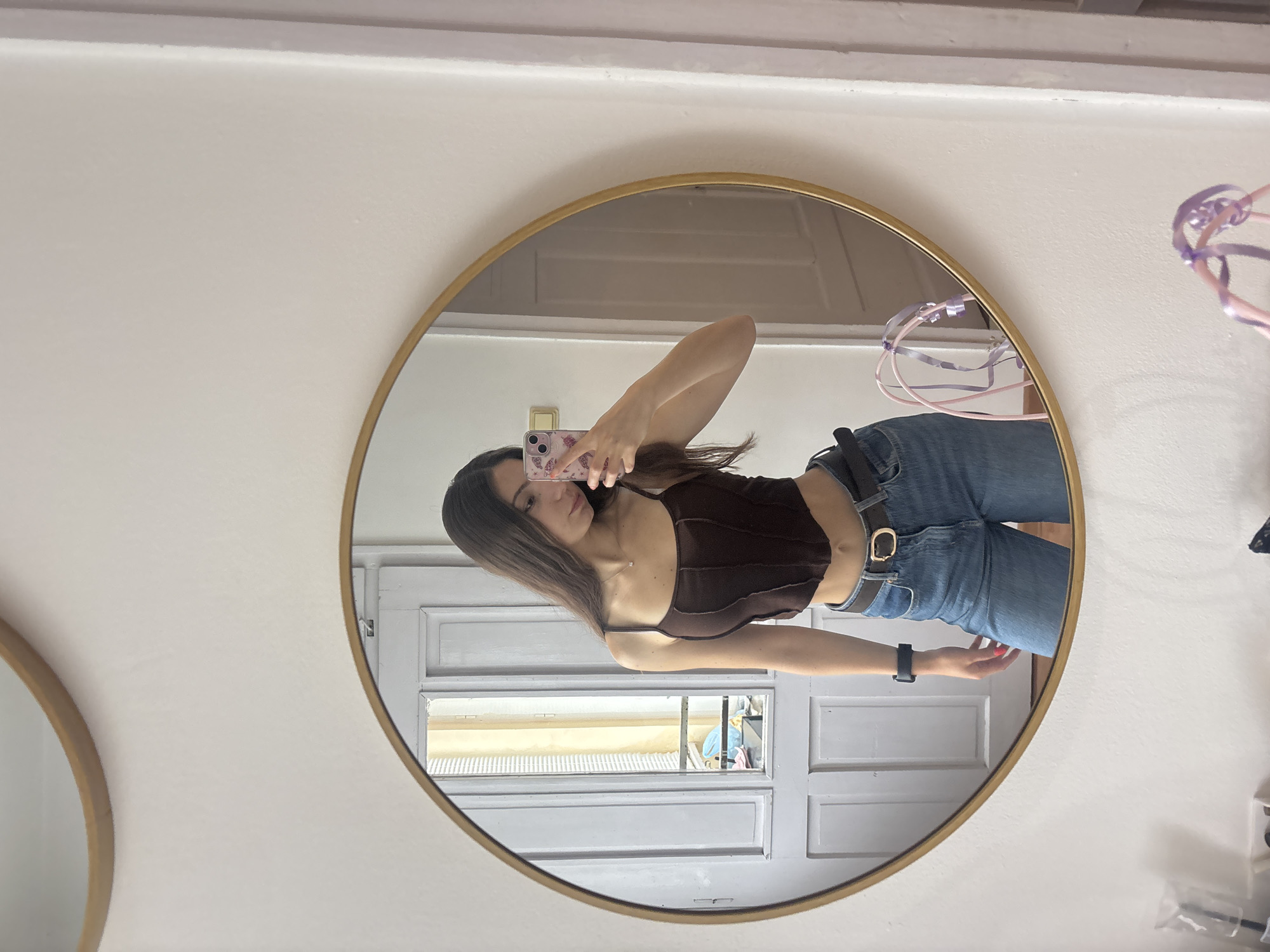 A young woman is taking a selfie in a round mirror with a gold frame. She is wearing a dark brown tank top and blue jeans with a belt. The background reflected in the mirror shows a white door with a window. A wall with another round mirror is visible on the left side of the image. There is a white wall on the right side. The location is not identifiable as Kaspichan, Bulgaria.