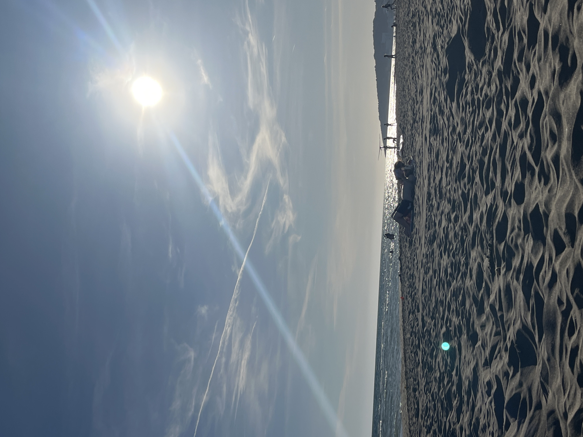 The image shows a beach scene in Donji Štoj, Montenegro, taken during bright daylight. The sky is blue with wispy clouds and a contrail from an airplane. The sun is high in the sky, with a strong glare. The beach is sandy, and there are people on the shoreline and in the distance. The sea is visible, reflecting the sunlight.
