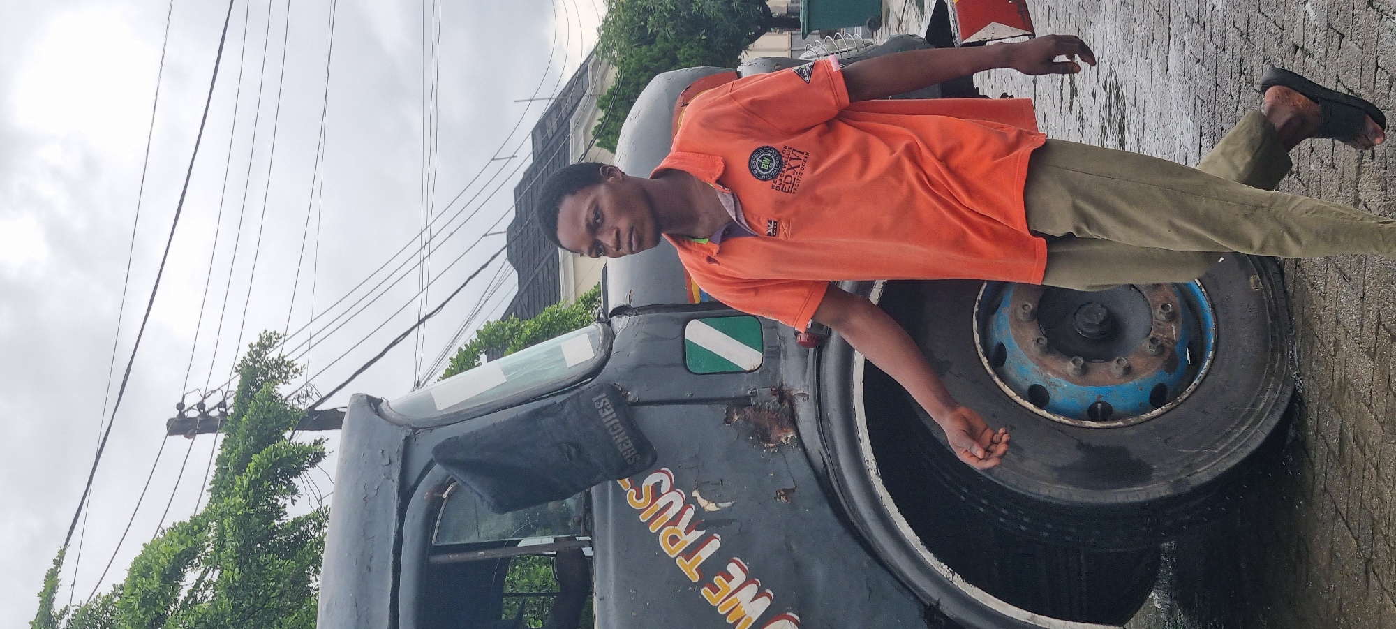 A young man walks beside a dark, weathered truck parked on a cobblestone street in Moba, Nigeria. The truck has white lettering that reads "WE TRUST" and a green and white emblem. The man wears an orange polo shirt and olive-colored trousers. The sky is overcast, and the ground appears wet, suggesting recent rain. In the background, a house with a dark roof and some trees are visible. Electrical wires crisscross the sky.