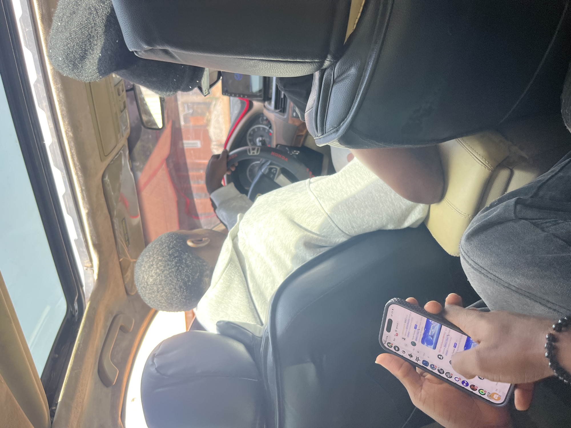 This media file depicts the interior of a vehicle, likely in Barakin Akawo, Nigeria. The scene is dominated by the seating and dashboard of the car. In the foreground, a person's hands are visible holding a smartphone, which displays various icons and text, possibly a social media or messaging application. The phone has a dark, patterned case. The person's fingers are interacting with the touchscreen.
Behind the hands, another individual is seated, facing away from the camera and towards the steering wheel. This person is wearing a light-colored hoodie and appears to be either driving or about to drive, with their hands on the steering wheel. The steering wheel has red accents and the "HONDA" logo is visible. A portion of the car's dashboard, including a screen and gauges, is also in view.
The interior of the car appears to be somewhat worn. The seats are upholstered in what looks like black leather and beige fabric. There are also fuzzy, dark-colored accessories attached to the interior of the car. The lighting suggests it is daytime, with natural light coming through the windows. The overall mood is casual and everyday.