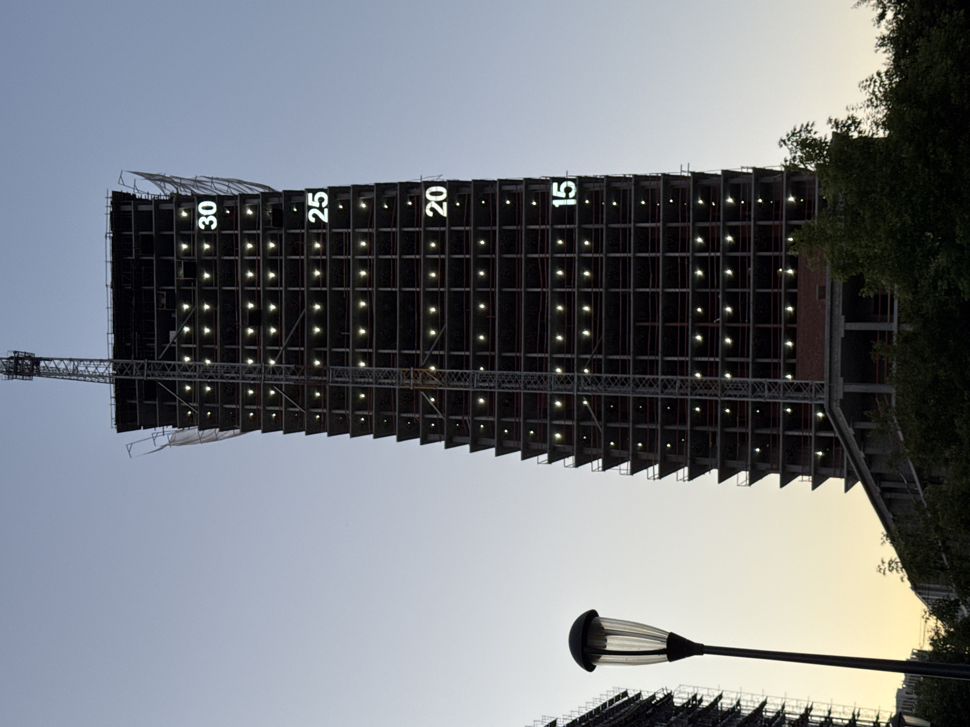 This image captures a construction site in Izhevsk, Russia, featuring a tall, unfinished skyscraper against a clear sky. The building's skeletal structure is visible, with numerous lights illuminating its floors. White numerals are projected onto the side of the building, indicating "30," "25," "20," and "15," suggesting floor numbers or a countdown. A construction crane is attached to the top of the skyscraper. In the foreground, a street lamp stands on the left side of the frame, with lush green trees and other buildings partially visible at the bottom. The scene appears to be set during dusk or early evening, given the sky's gradient and the illuminated lights.