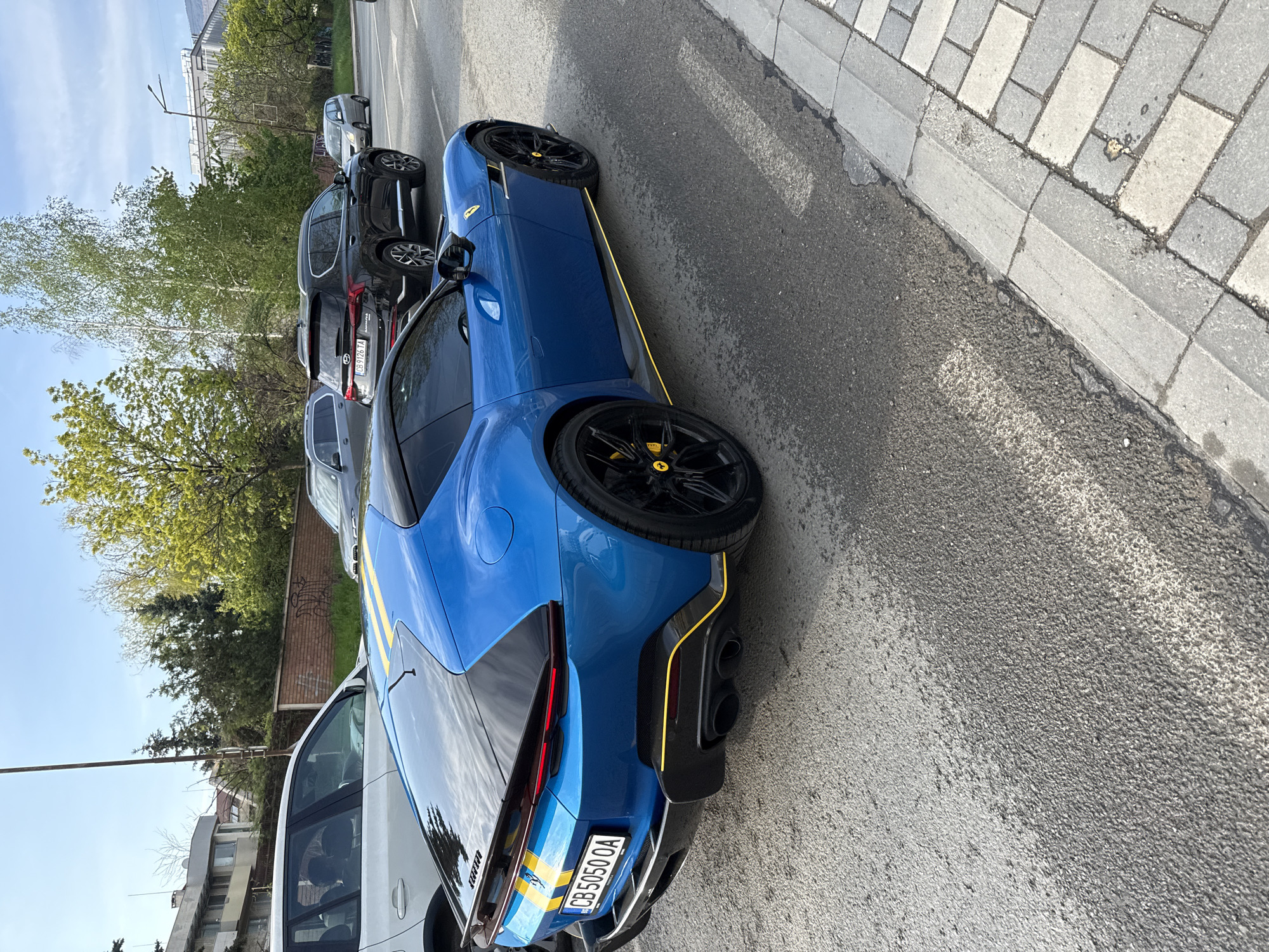 A vibrant blue Ferrari, likely a Roma, is prominently parked on the right side of an asphalt street in Sofia, Bulgaria. The car features distinctive yellow accents on its side sills, rear diffuser, and subtle stripes on the rear bumper. Its black multi-spoke wheels house bright yellow brake calipers, and the wheel center caps bear the Ferrari logo. The Bulgarian license plate "CB 5050 OA" is clearly visible on the rear, with "CB" signifying Sofia.
Behind the Ferrari, several other cars are parked, including a dark SUV with the Bulgarian license plate "CB 9126 TA" and a grey vehicle. In front of the Ferrari, partially visible, is a white car, and further down the street, another light-colored car is parked.
The scene takes place on a bright, sunny day, indicated by clear blue skies and strong shadows. Lush green trees with fresh spring foliage line the side of the road, suggesting the season is spring or early summer. A grey paved sidewalk runs parallel to the street on the right. In the background, various buildings are visible under the clear sky. No people are present, and no activity other than parked vehicles is depicted. The word "Ferrari" is reflected on the side mirror of the white car next to the blue sports car.