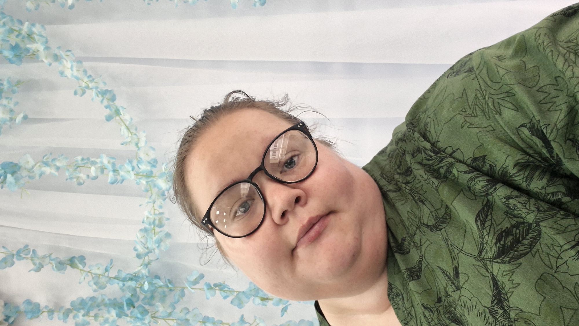 A close-up, tilted portrait features a person with fair skin, glasses, and dark hair pulled back, looking directly at the camera with a neutral expression. The person wears a dark green garment adorned with a black outline pattern of tropical leaves and flora. The black-rimmed glasses have reflections in their lenses, showing what appears to be a window or a white interior structure with horizontal divisions.
The background is a light, possibly white or pale blue, surface with horizontal folds or stripes. Draped across this background are several strands of artificial light blue and white flowers, resembling wisteria or similar cascading blossoms. The overall setup suggests an indoor setting, possibly with a decorative backdrop. The image is significantly rotated counter-clockwise. There is no visible text. The scene is located in Chaikovsky, Russia.