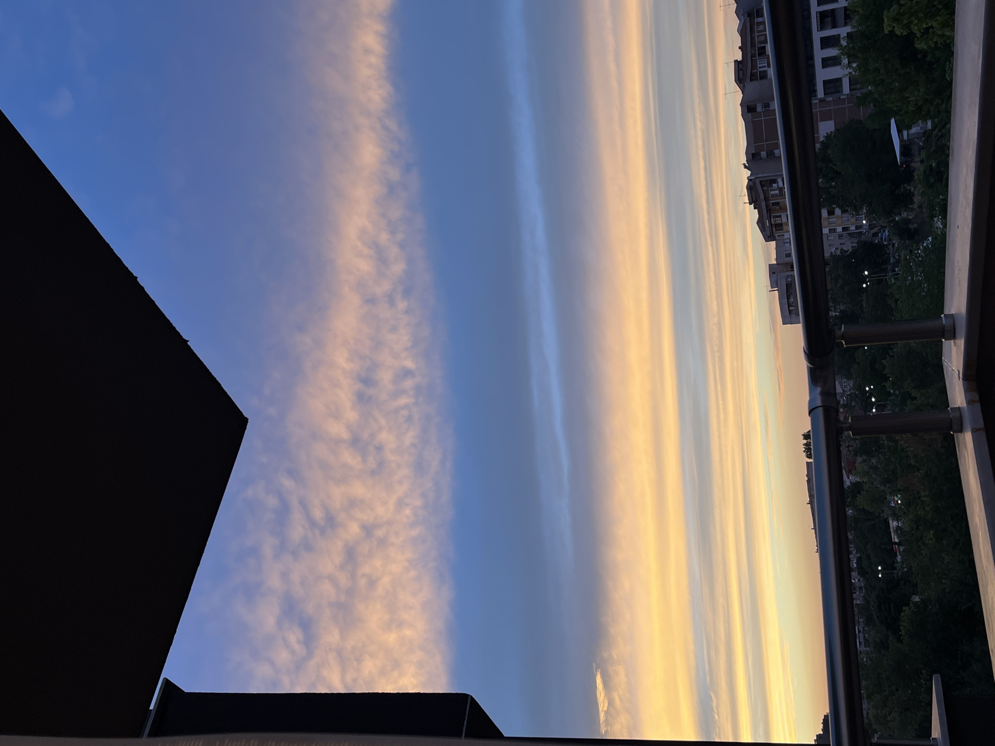 The image captures a sunset view from a balcony in Plovdiv, Bulgaria. The sky is a vibrant gradient of colors, with streaks of pink and orange clouds against a deepening blue. Below the sky, a row of apartment buildings is visible, interspersed with lush green trees. A metal railing runs across the foreground, partially obscuring the view of the city below. The scene appears tranquil, with no people or activity evident. The time of day is clearly evening, as indicated by the sunset hues and the streetlights beginning to illuminate the city.