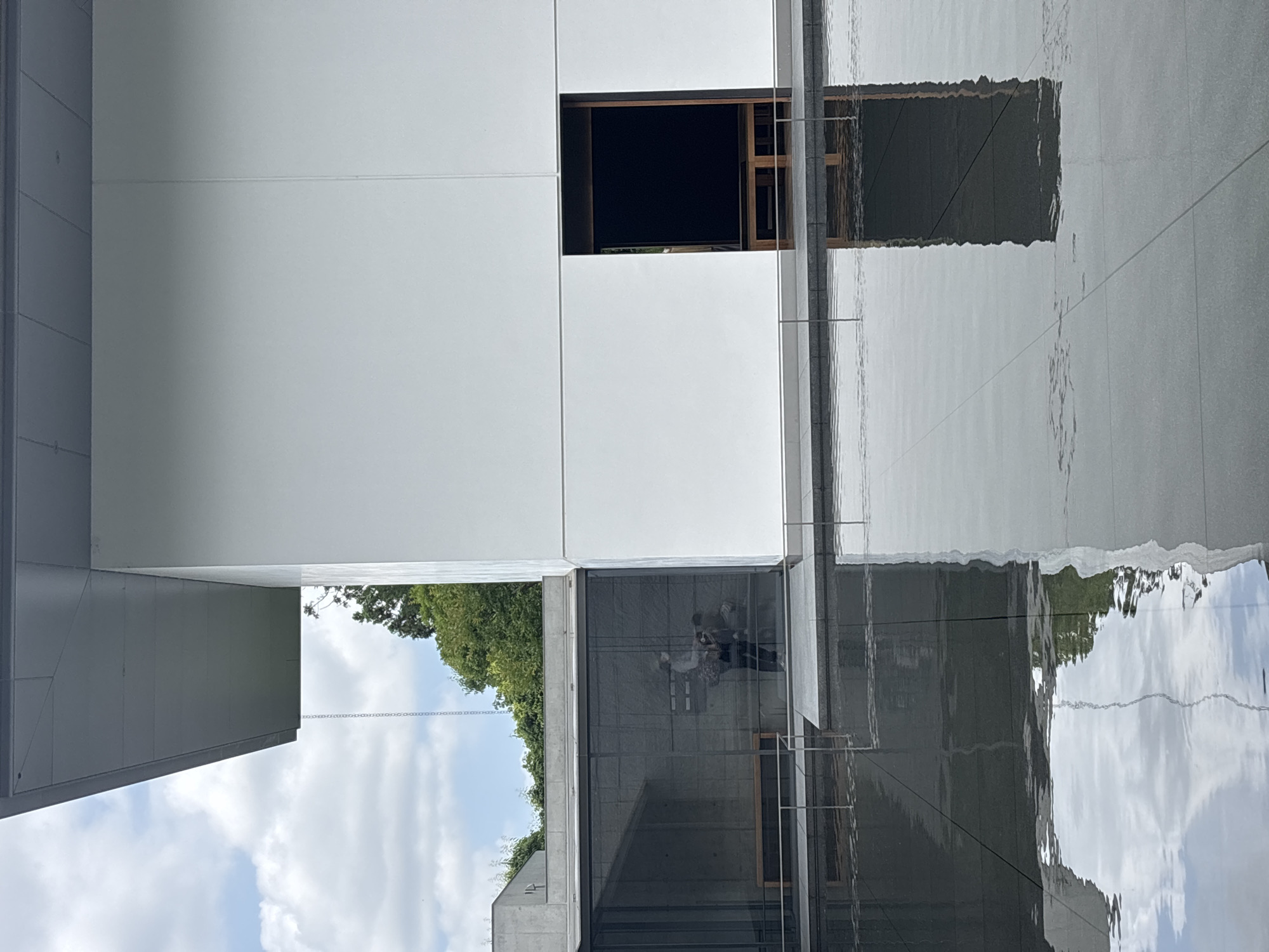 The image depicts a modern architectural complex at the 21st Century Museum of Contemporary Art in Kanazawa, Japan, viewed across a large, dark-tiled water feature that occupies the foreground.
The building on the right features a stark, multi-paneled white facade with clean horizontal lines. A large, dark, rectangular opening, possibly an entrance or deep recess, is present in the lower right section of this facade, revealing a dark interior with visible light brown wooden elements at its base.
To the left of the white facade, a dark-tinted glass wall extends across the mid-ground. This glass wall has a visible grid structure and reflects the interior, where at least two human figures are discernible. One figure, wearing a light-colored top and a face mask, is centrally reflected. Faint outlines of what appear to be light brown benches are also reflected inside. Beyond the glass wall, another section of the building with a flat, light grey roof is visible, with green trees peaking over it and in the distance.
A dark grey, flat roof overhang extends from the top left of the building against a partly cloudy blue sky. A slender, silver-colored metal rain chain hangs vertically from the edge of this overhang, disappearing towards the water feature.
A thin, grey metal handrailing runs horizontally along the edge between the building's base and the water feature. The water itself is dark and reflective, mirroring the sky with patches of white clouds and the building's structures, including the white facade, the dark opening, and the rain chain which creates a distinct wavy vertical reflection. The bottom of the water feature reveals a grid of large, dark grey square tiles. The water's surface exhibits ripples, distorting the reflections.