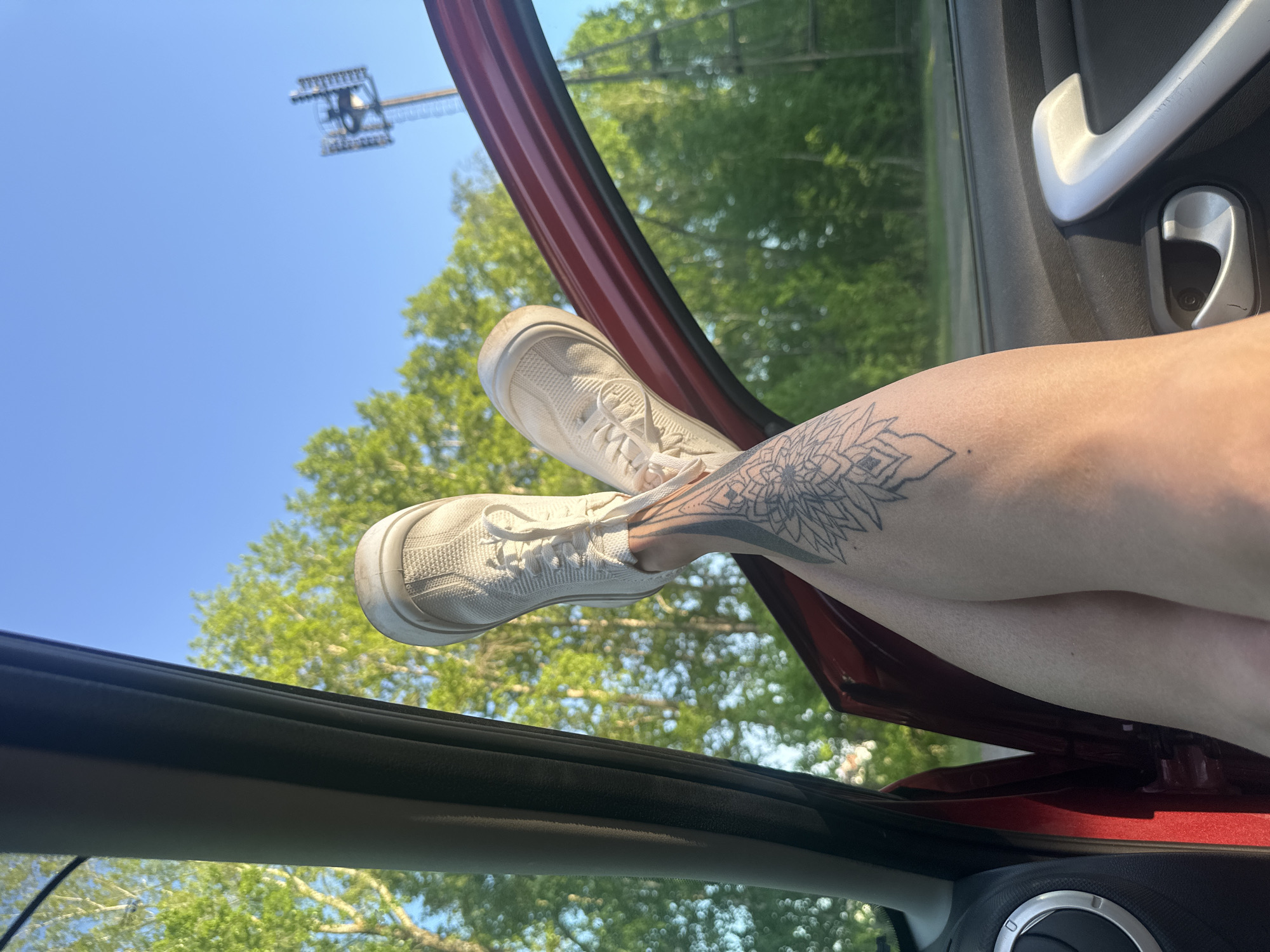 The image shows a view from inside a red car, with a person's legs and feet sticking out of the open car door. The person is wearing white sneakers, and there is a tattoo on their leg. Through the car door, there are trees, a blue sky, and a tall metal structure with a crane. It is unclear if this image location is Chaikovsky, Russia.