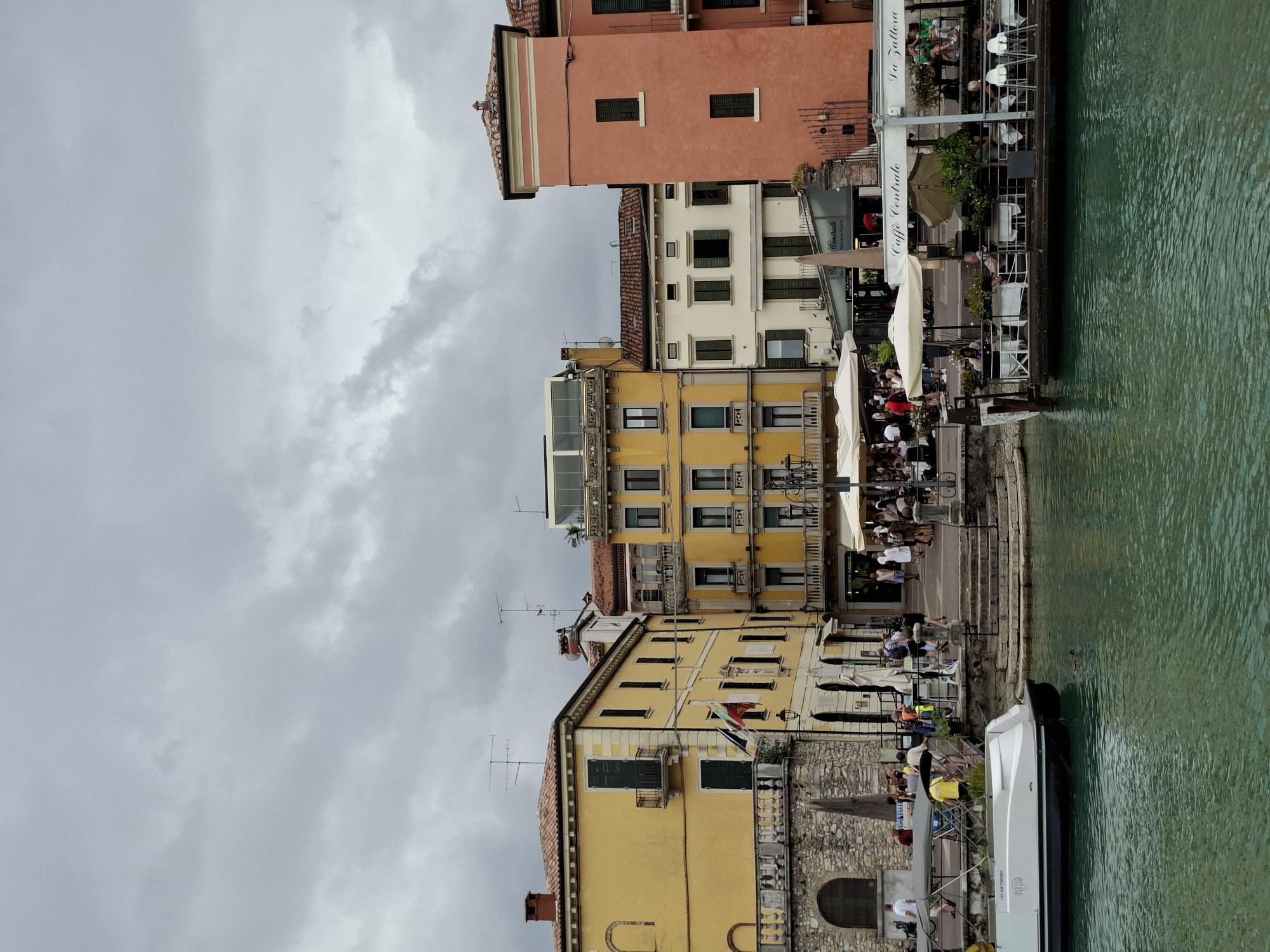 This image depicts a cloudy day in Peschiera del Garda, Italy, specifically along its waterfront. The scene is dominated by several multi-story buildings with distinct architectural styles. The building on the far left is a yellowish-ochre color with a red-tiled roof and a stone lower level. It features arched windows and a balcony. Next to it is another yellow building, taller and more ornate, with balconies and numerous windows. To the right, a white building with green shutters is situated between the yellow buildings and a vibrant, salmon-pink building on the far right, which also has green shutters.
In the foreground, a bright turquoise body of water is visible. A white pontoon boat with a canopy is docked on the left side of the frame, and a smaller duck is swimming in the water. On the right side, a floating restaurant with white umbrellas and outdoor seating is partially visible, with patrons seated at tables. The restaurant appears to have signage that reads "Caffe Centrale" and "La Zattera".
Along the waterfront, a walkway is lined with more umbrellas and tables from outdoor cafes. People are seen walking along the promenade, some standing near the water's edge, and others seated at the cafes. Steps lead down from the promenade to the water's edge in the center of the image. Flags are visible hanging from one of the buildings. Antennas are mounted on the roofs of some of the buildings. The overall atmosphere is that of a bustling, European waterfront town on an overcast day.