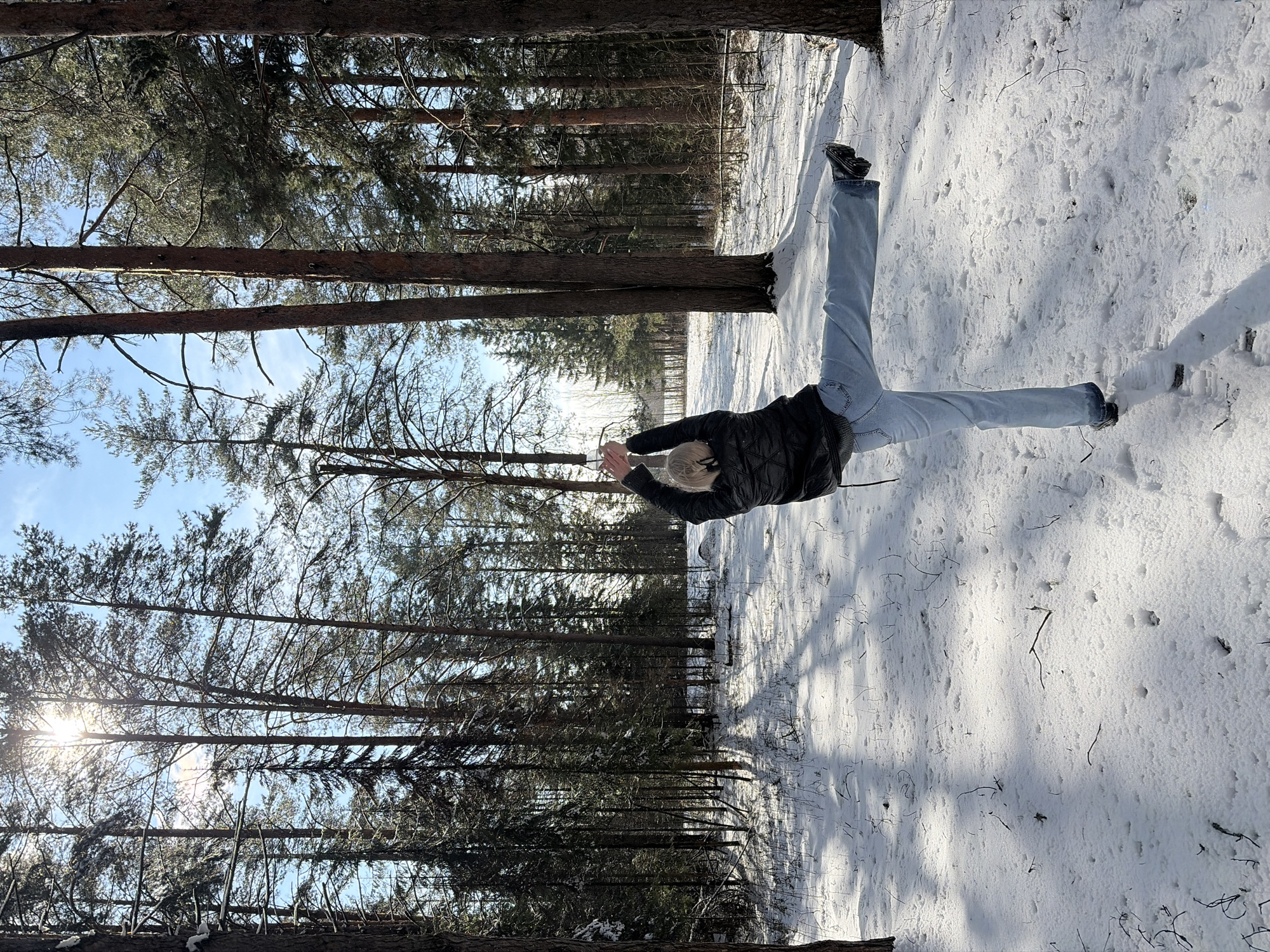 The photograph captures a winter scene in what appears to be a forest or wooded area, potentially near Прикамский, Russia. The ground is covered in snow, and the image is filled with tall, thin trees. A person is performing a split, their legs extended in opposite directions, with their body positioned horizontally. The person is wearing jeans, a black jacket, and dark footwear. The sun is visible through the trees, creating shadows and highlighting the snowy landscape.