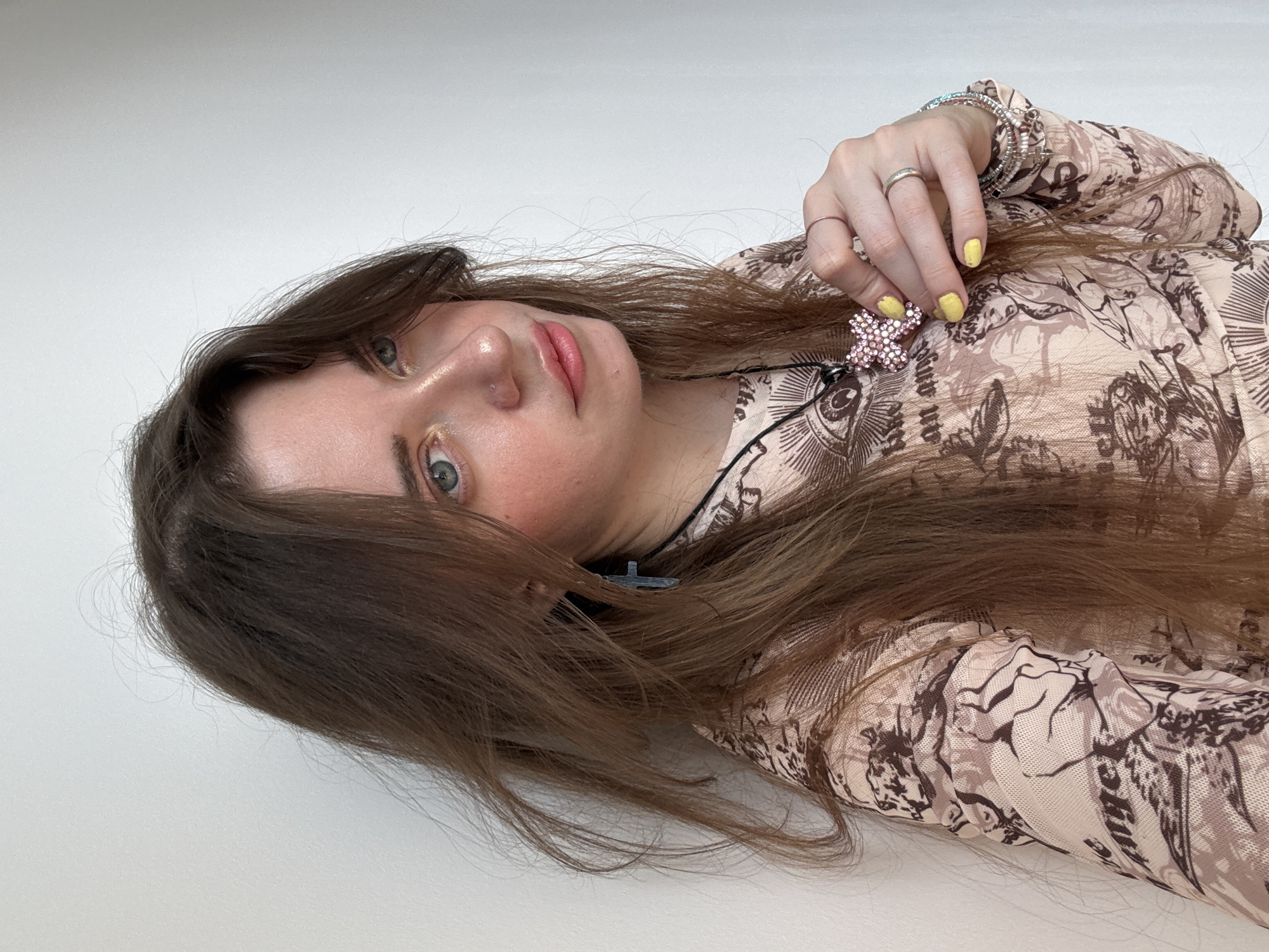 A young woman with long brown hair and green eyes is lying on a white surface. She is wearing a patterned beige shirt. Her fingernails are painted yellow. She is holding a pink and yellow cross-shaped pendant. She is wearing a ring on her finger and bracelets on her wrist. A black cord with a silver pendant is visible around her neck. The location of the image is not discernible.