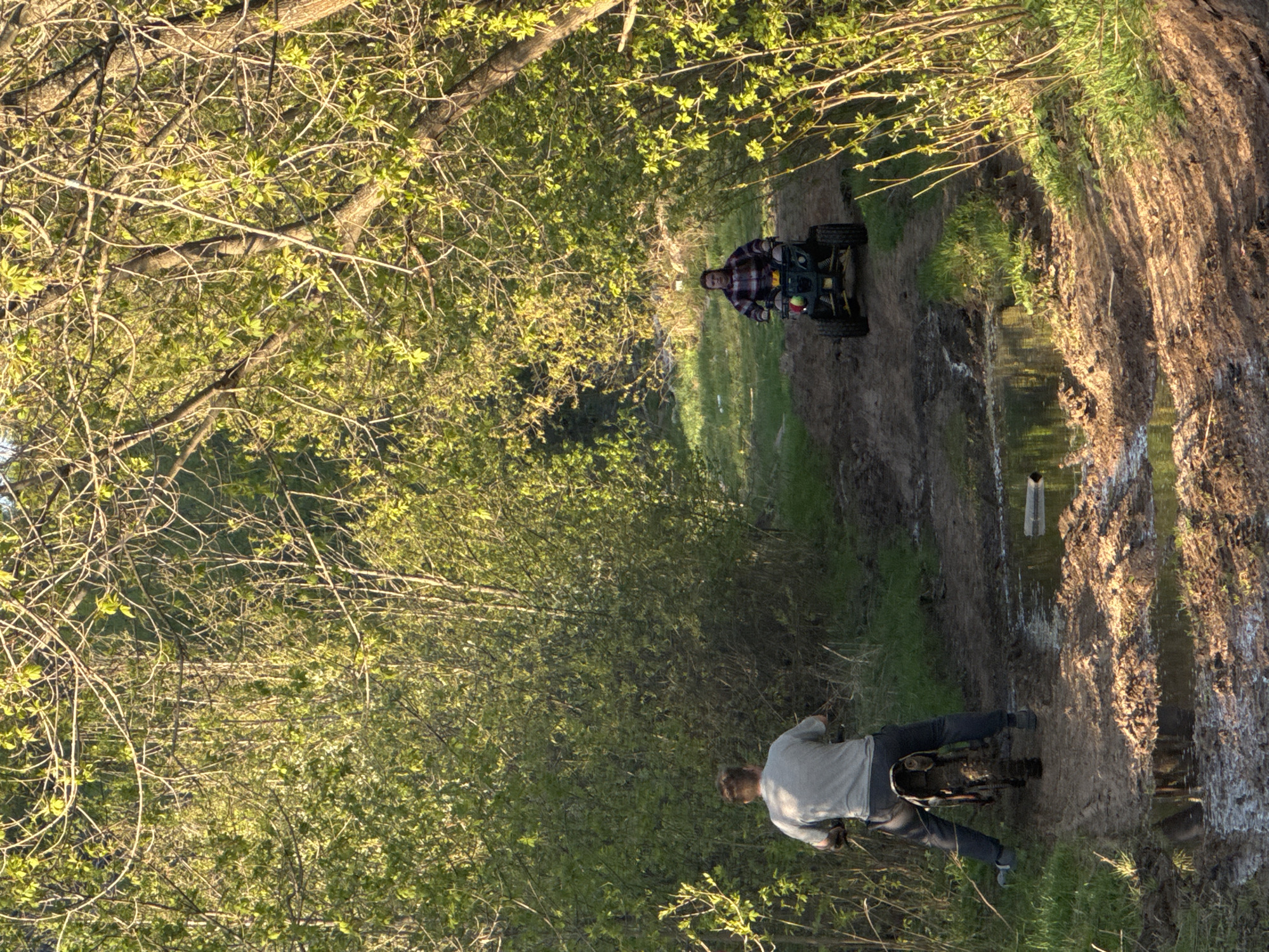 The image shows a dirt path through a wooded area. A man is riding a motorcycle on the path in the foreground. Further down the path, another man is riding a four-wheeled vehicle. The path is muddy with standing water. The trees are dense with green leaves. The scene is located in Perm, Russia.