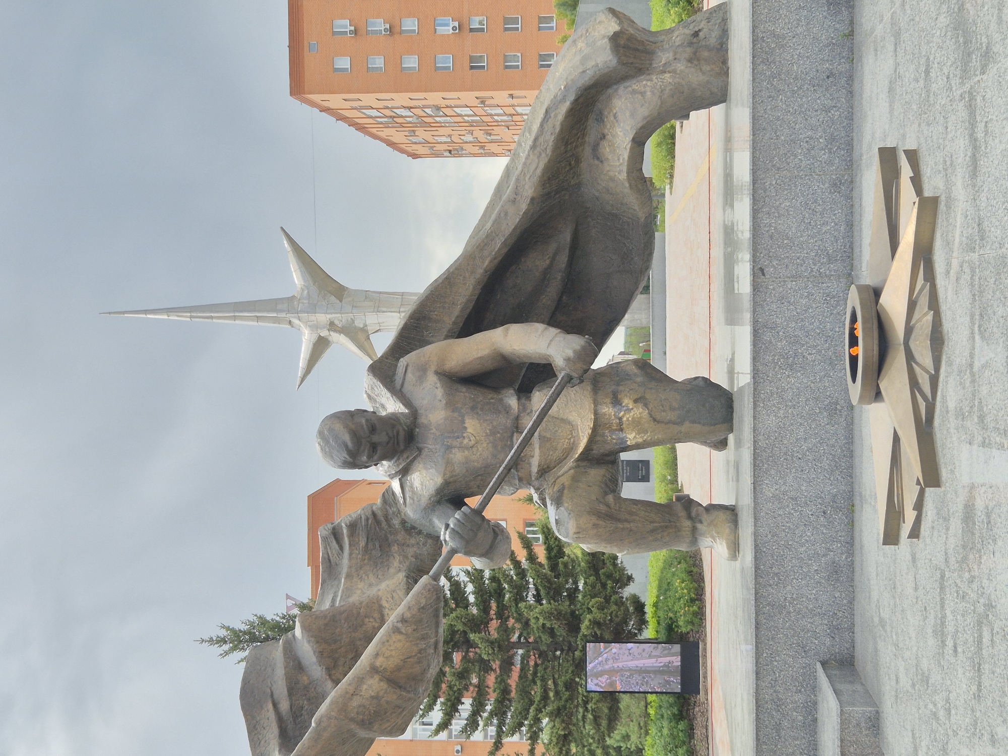 The image depicts a war memorial in Kokshetau, Kazakhstan. In the foreground, a bronze sculpture of a kneeling soldier holding a flag is prominently displayed. The soldier is in military attire, and the flag appears to be a significant part of the monument, draped around him. In front of the sculpture, a perpetual flame burns within a star-shaped structure, symbolizing remembrance. Behind the sculpture, a tall, silver, star-shaped monument rises against a cloudy sky. Two modern, multi-story residential buildings, typical of Soviet-era architecture, flank the memorial. Green trees and manicured bushes surround the monument, suggesting a well-maintained public space. A digital display screen is visible to the left of the main sculpture, showing an image of people gathered in a procession. The overall scene conveys a sense of solemnity and historical significance, commemorating a past event.