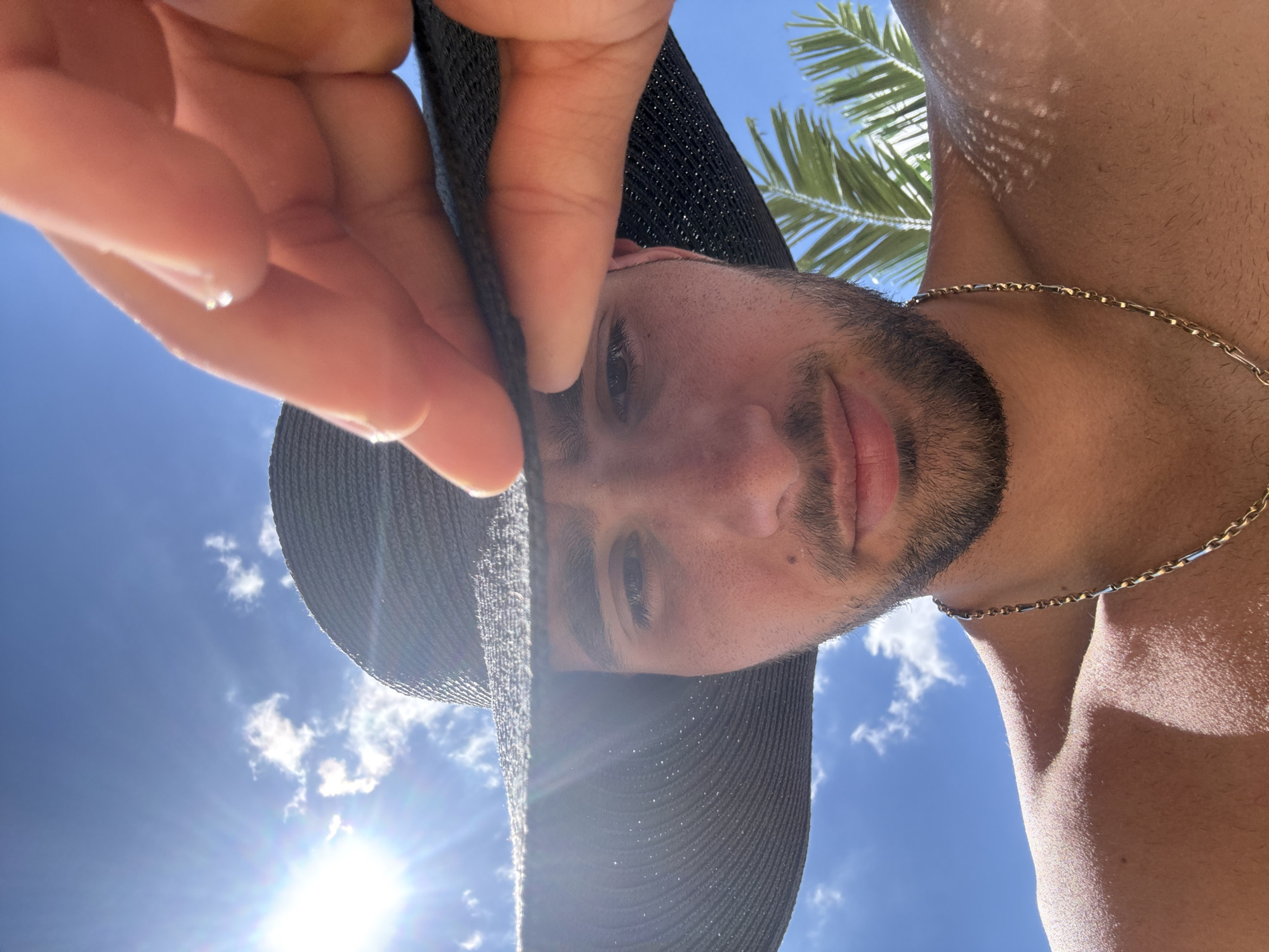 A man wearing a wide-brimmed black hat and a gold chain is looking up at the camera, partially obscuring his face with his hand. He has a beard and is shirtless. The background is a bright blue sky with some clouds and sunlight, and palm fronds are visible on the right side of the image. The scene appears to be outdoors in a sunny location, possibly Nesebar, Bulgaria, given the presence of palm trees and bright sunlight.