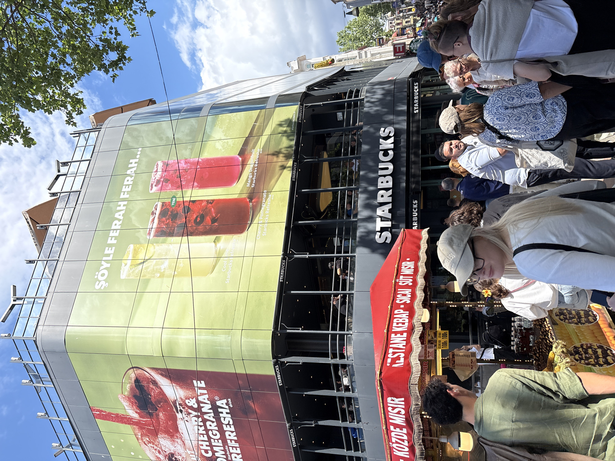 Here's a description of the image:
The image depicts a bustling urban scene, likely taken during the day in Kadıköy, Turkey. A large, multi-story Starbucks building is prominent, its facade covered in advertisements for "Starbucks Refresha" drinks, featuring images of cherry and pomegranate, mango dragonfruit, and other flavors. The text on the advertisement is partially in Turkish ("Şöyle Ferah Ferah...").
A crowd of people is gathered along the sidewalk in front of the Starbucks. Closer to the camera, a vendor's stall or stand is visible, indicated by a red awning with Turkish text advertising "Közde Mısır" (grilled corn) and "Kestane Kebap - Sıcak Süt Mısır" (chestnut kebab - hot milk corn). People appear to be interacting with the vendor, possibly making purchases.
Several people in the crowd are partially visible, including individuals wearing casual clothing, hats, and sunglasses. The overall environment suggests a busy, commercial area within Kadıköy, potentially a popular pedestrian zone.