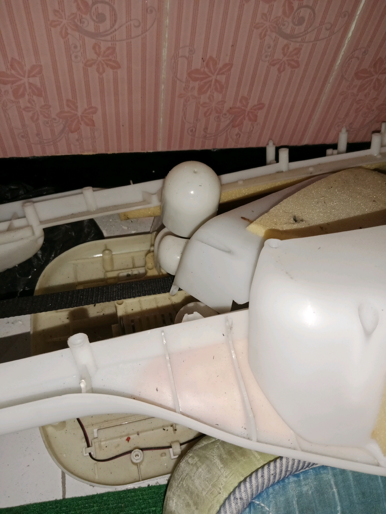 The image displays a close-up, overhead view of the internal components of a device, likely a washing machine. The scene is dominated by white plastic structures, some of which appear to be housing or guiding mechanisms. A black rubber belt is visible stretched across a portion of the device. There are also pieces of foam material present, one of which is a pale yellow and has a rough texture. The upper portion of the image shows a pink-patterned surface, possibly wallpaper or a decorative covering, with vertical stripes and floral motifs. The scene is cluttered and appears to be in a state of disrepair or disassembly, with dust and debris visible on some surfaces. There are no people or animals in the image. The location is specified as Ringinrejo, Indonesia.