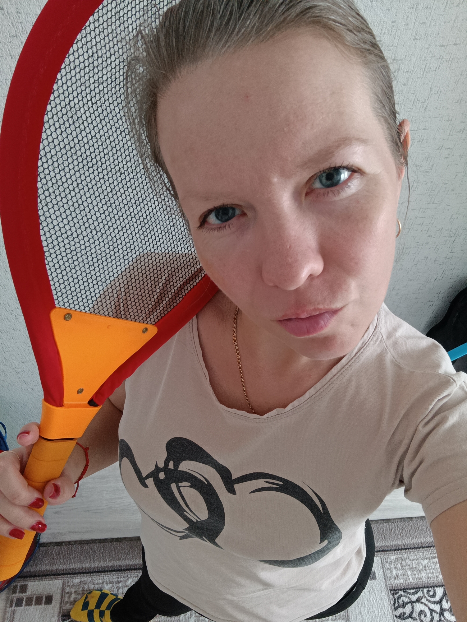 A woman is holding a badminton racket in a room with wallpapered walls. She has fair skin, blue eyes, and her hair is pulled back. She's wearing a beige t-shirt with a black design, a gold necklace, and earrings. Her fingernails are painted red. The badminton racket is red, orange, and yellow. The background includes part of the floor with a gray and beige patterned rug. The photograph appears to be taken from above. No specific location in Chaikovsky, Russia, can be determined from the image.