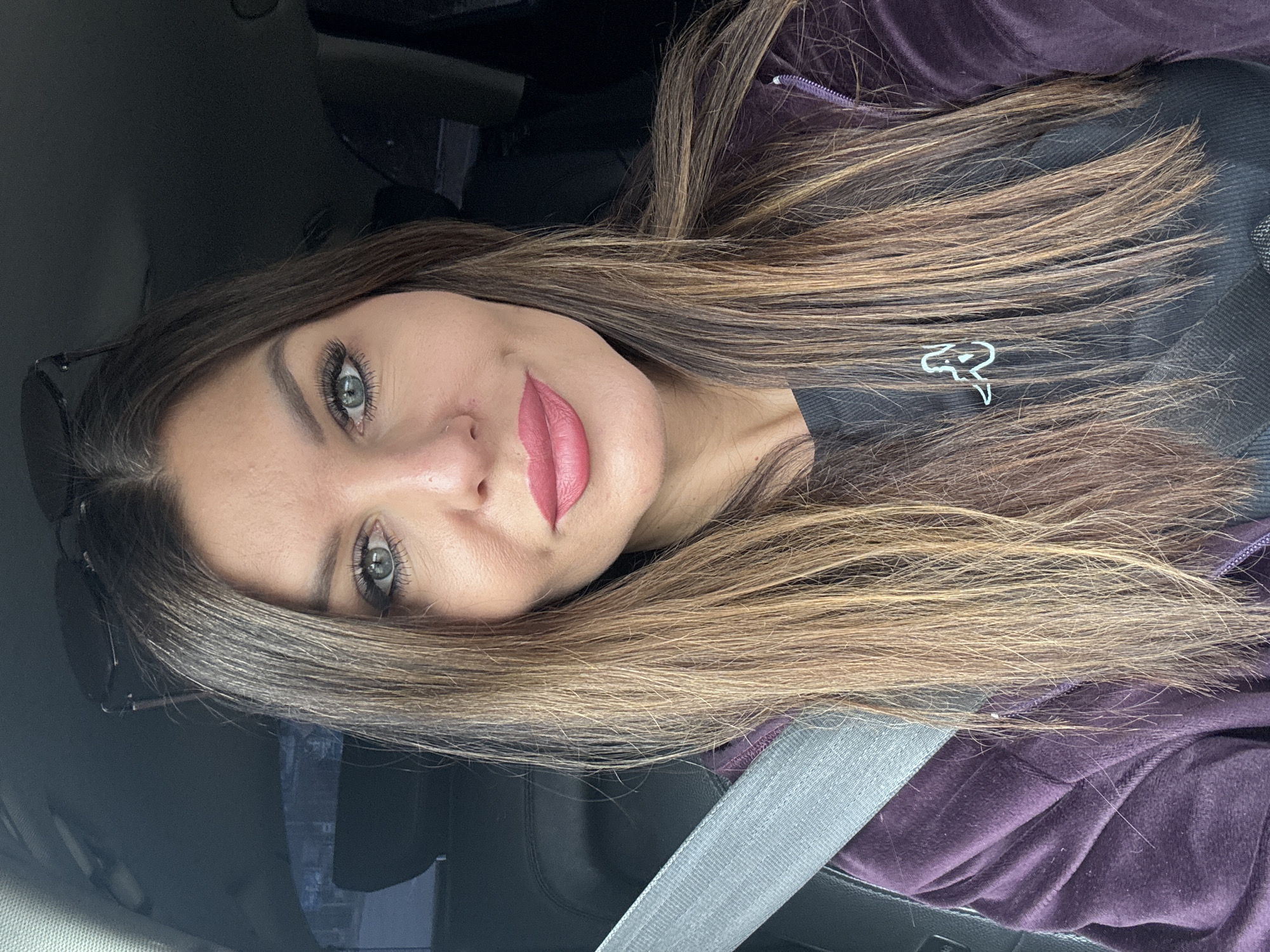 A medium-shot selfie of a woman with long brown hair and hazel eyes, likely taken in a car. She's wearing a dark t-shirt with a small white logo on the chest, under a purple zip-up hoodie. A silver seatbelt is across her chest. The background is dark, suggesting the car's interior. No specific location or evidence indicates the scene is in the city of Chaikovsky, Russia.