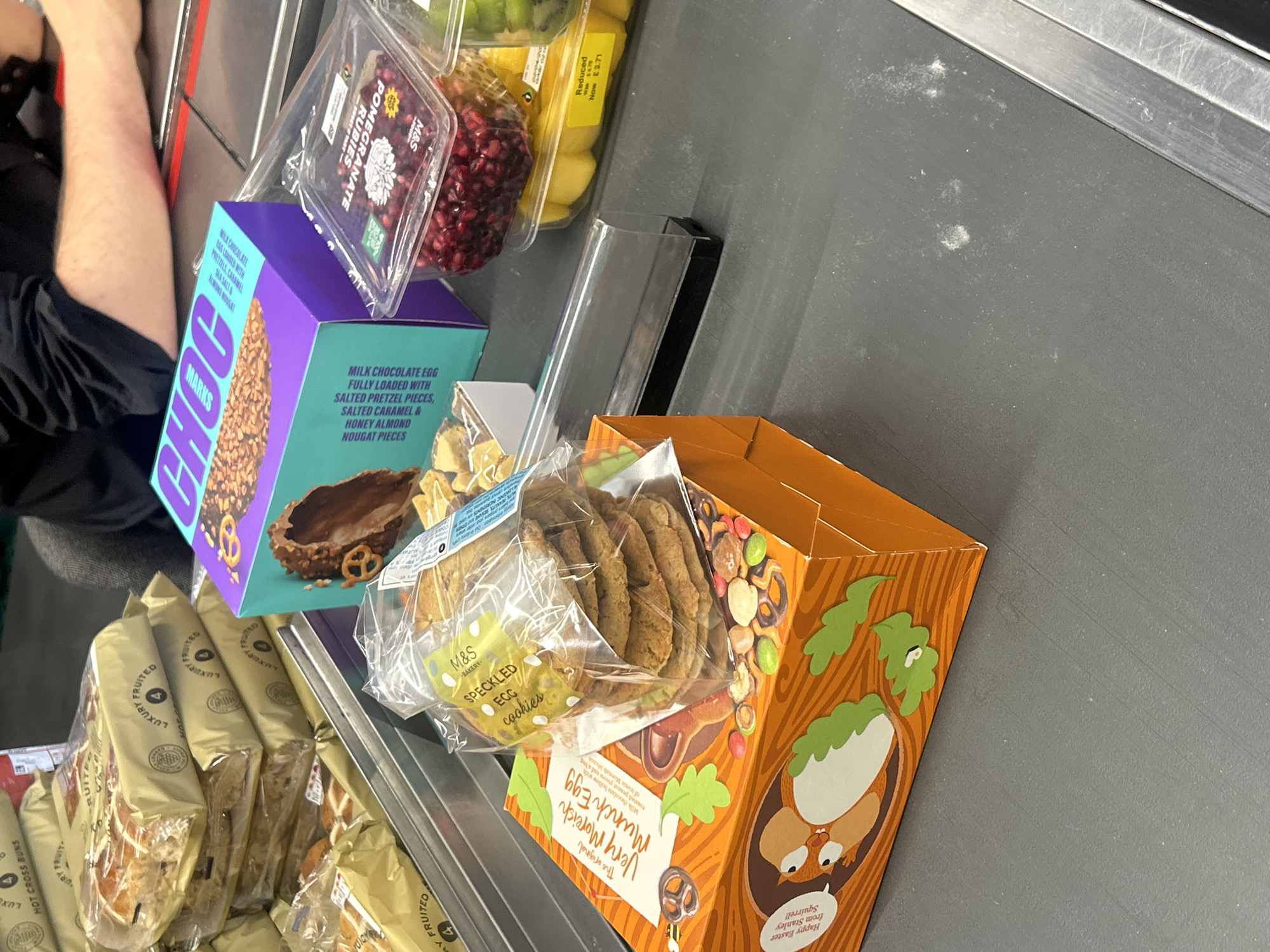 The image depicts a selection of grocery items on a checkout conveyor belt, likely in a supermarket located in Chippenham, United Kingdom, based on the branding of the products.
Visible items include:
- "LUXURY FRUITED HOT CROSS BUNS"
- "MARKS CHOC"
- "M&S SPECKLED EGG COOKIES"
- "Very Moreish Munch Egg"
The items are arranged close together and are partially obscuring each other, suggesting a recent placement on the conveyor belt in preparation for scanning at checkout.
The conveyor belt is grey, and a portion of a person's arm is visible.