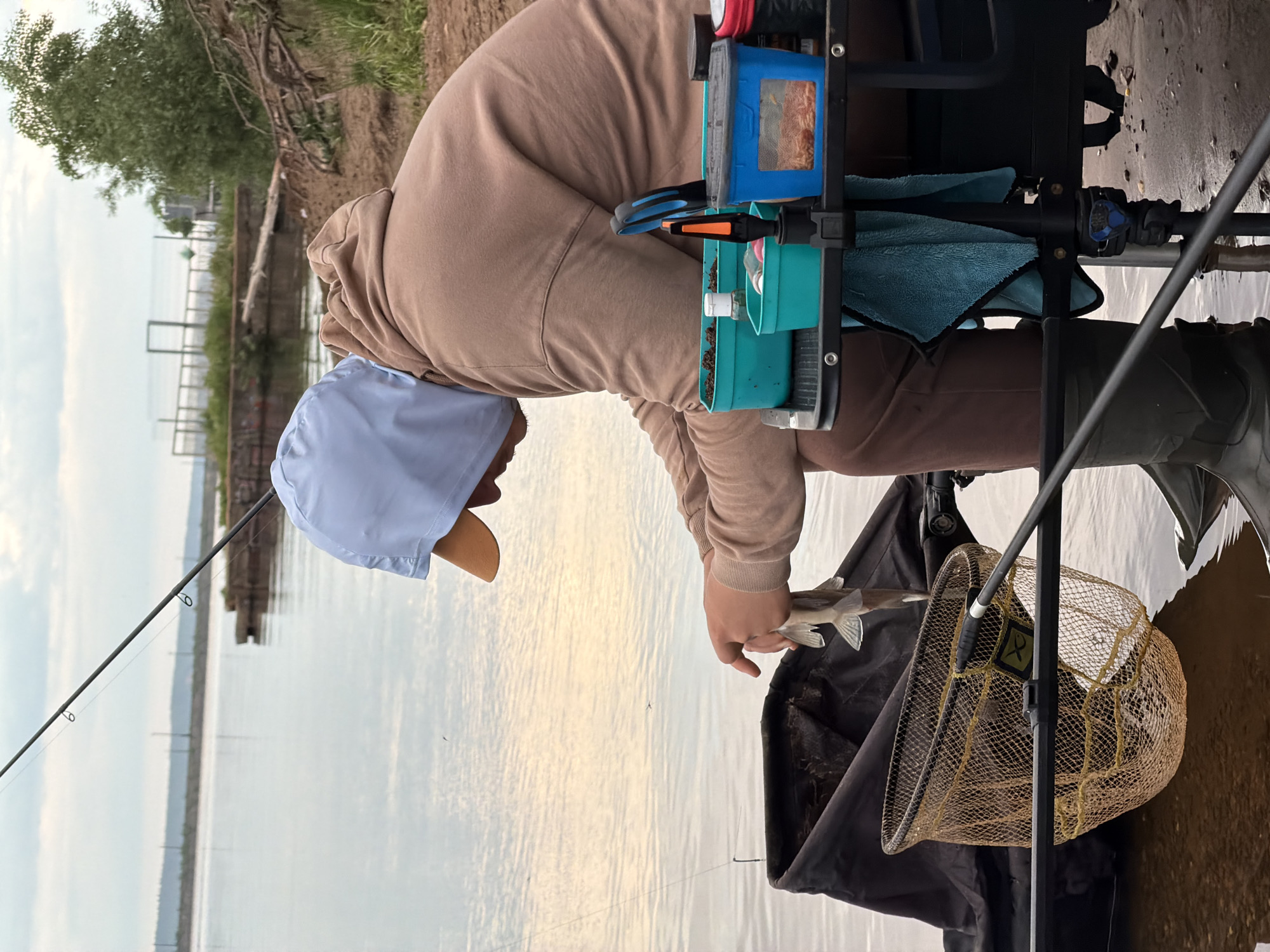 A person wearing a light brown hooded sweatshirt and a light blue hat is holding a small fish over a fishing net. The person is positioned near the edge of a body of water, likely a river or lake, as a fishing rod extends into the water on the left side of the frame. The location is Chaikovsky, Russia.
Behind the person, a fishing station setup is visible, including compartments and fishing gear. The ground near the water's edge is a mix of sand and wet areas. The backdrop features a blurred cityscape, suggesting the location is near an urban area. The sky is overcast, indicating a cloudy day.