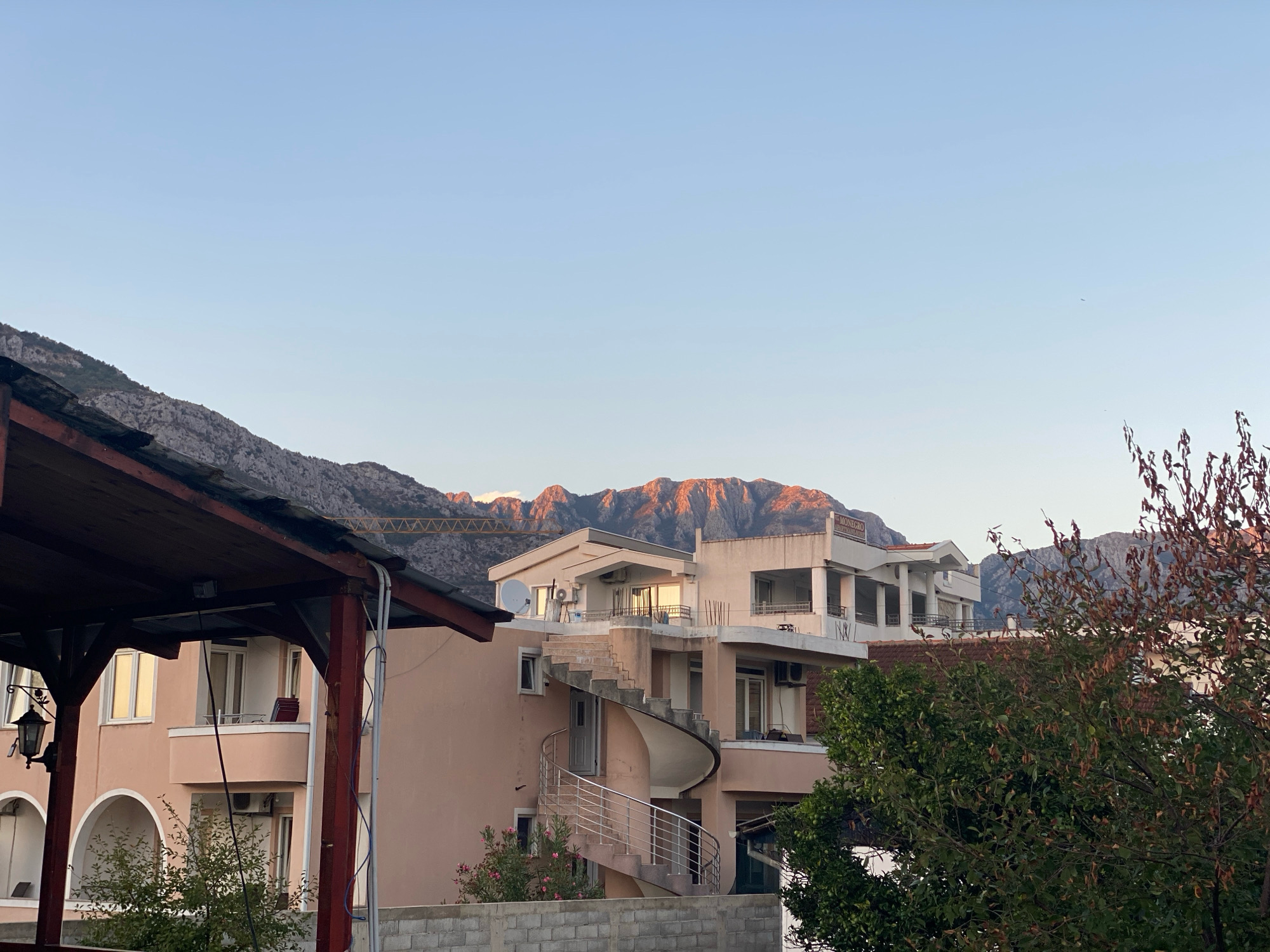 The image shows a view of buildings and mountains in the city of Bar, Montenegro, likely during the late afternoon or early evening. In the foreground, a wooden canopy structure partially obscures the view of a peach-colored building with arched doorways. To the right, a spiral staircase is visible on another building, leading to balconies. In the background, rugged mountains are illuminated by the setting sun, casting a warm glow on their peaks. A sign on one of the buildings reads "MONEGRO APARTMENTS". Trees with green and brown leaves are also present, suggesting it is autumn. The sky is a clear, pale blue.