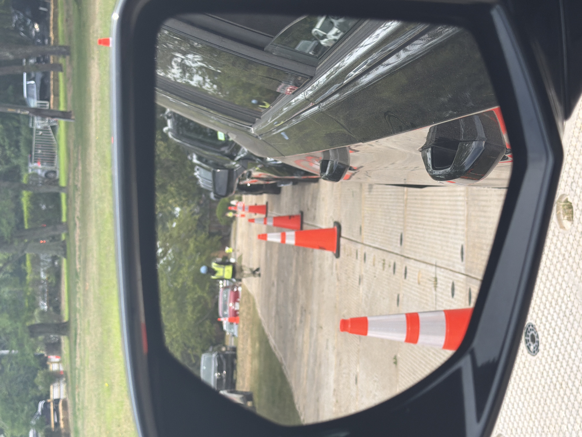 The image is a close-up view of a car's side mirror, reflecting a scene outdoors. In the reflection, a line of orange and white traffic cones is visible, marking a path. Behind the cones, several cars are parked or in a queue. Further in the background, a person wearing a bright yellow vest and a blue hard hat is walking. The environment appears to be a grassy area with trees, suggesting an outdoor event or gathering. The scene is set in the Town of Southampton, United States of America.