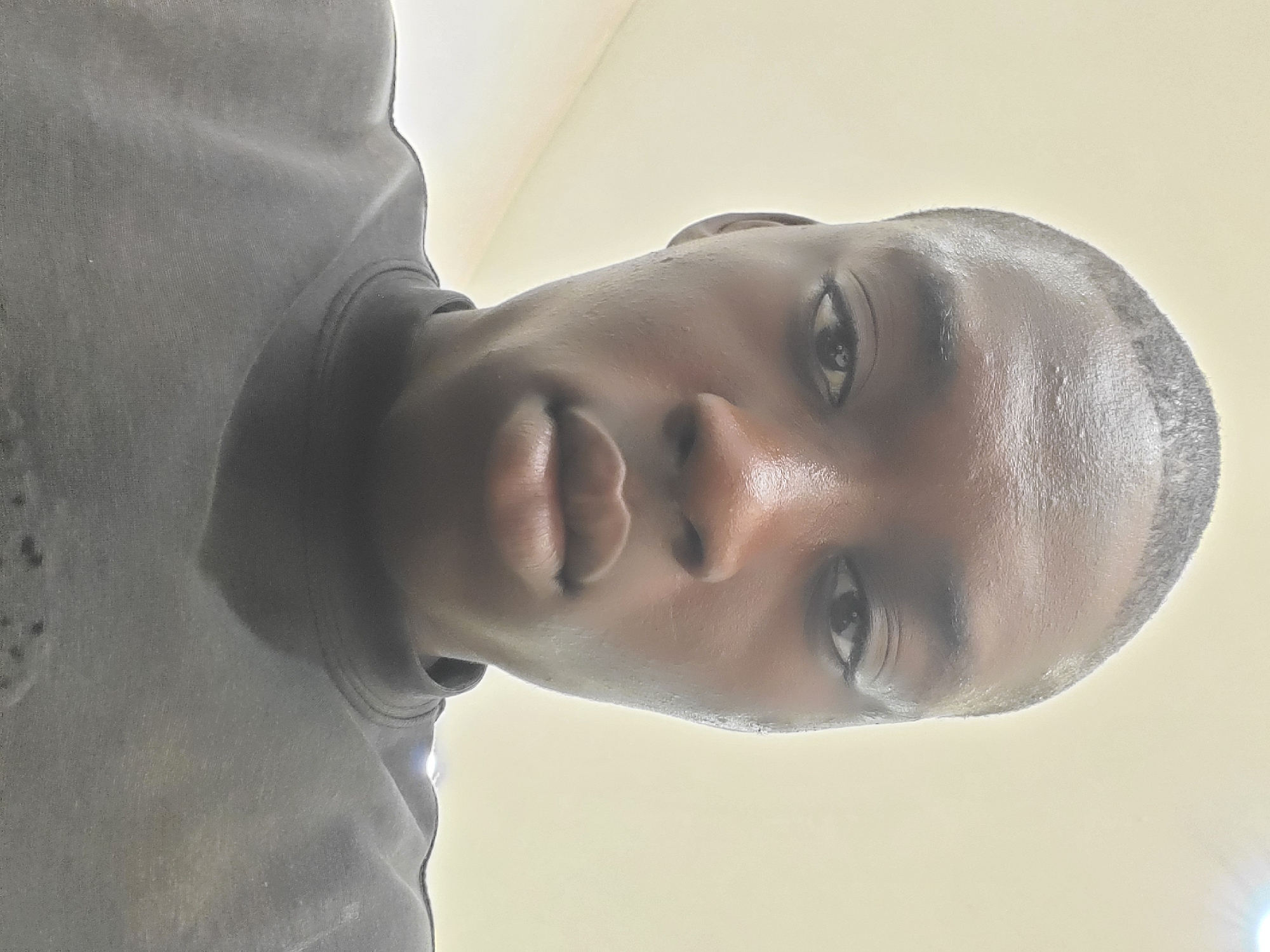 A medium close-up, low-angle shot depicts a dark-skinned person with a closely shaved head. The person's face is slightly shiny, possibly due to perspiration or skin oils. They are wearing a dark-colored sweatshirt. The background is a plain, light yellow or off-white surface. There is no information in the image to indicate the location is Fegge, Nigeria. The person's actions and interactions cannot be determined from the image. The environment is limited to the immediate background.