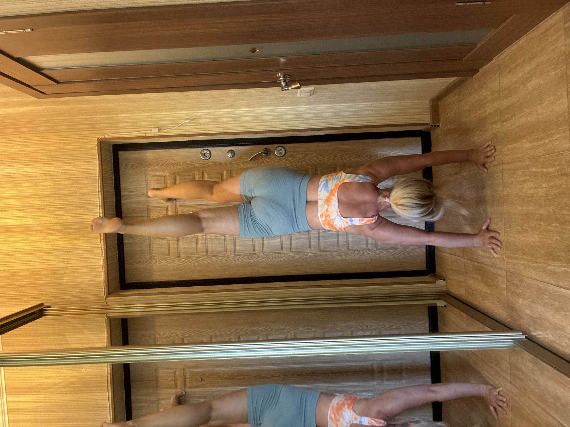 A woman with blonde hair tied in a ponytail is performing a handstand in a hallway. She is wearing a colorful sports bra and light blue shorts. Her left leg is extended upwards, while her right leg is bent at the knee. The hallway has light-colored, wood-grained wallpaper and tiled flooring. There is a large mirrored wardrobe on the left and two wooden doors on the right. The overall scene suggests an indoor setting, possibly a residential hallway. There is no information to suggest this image was taken in Sochi, Russia.