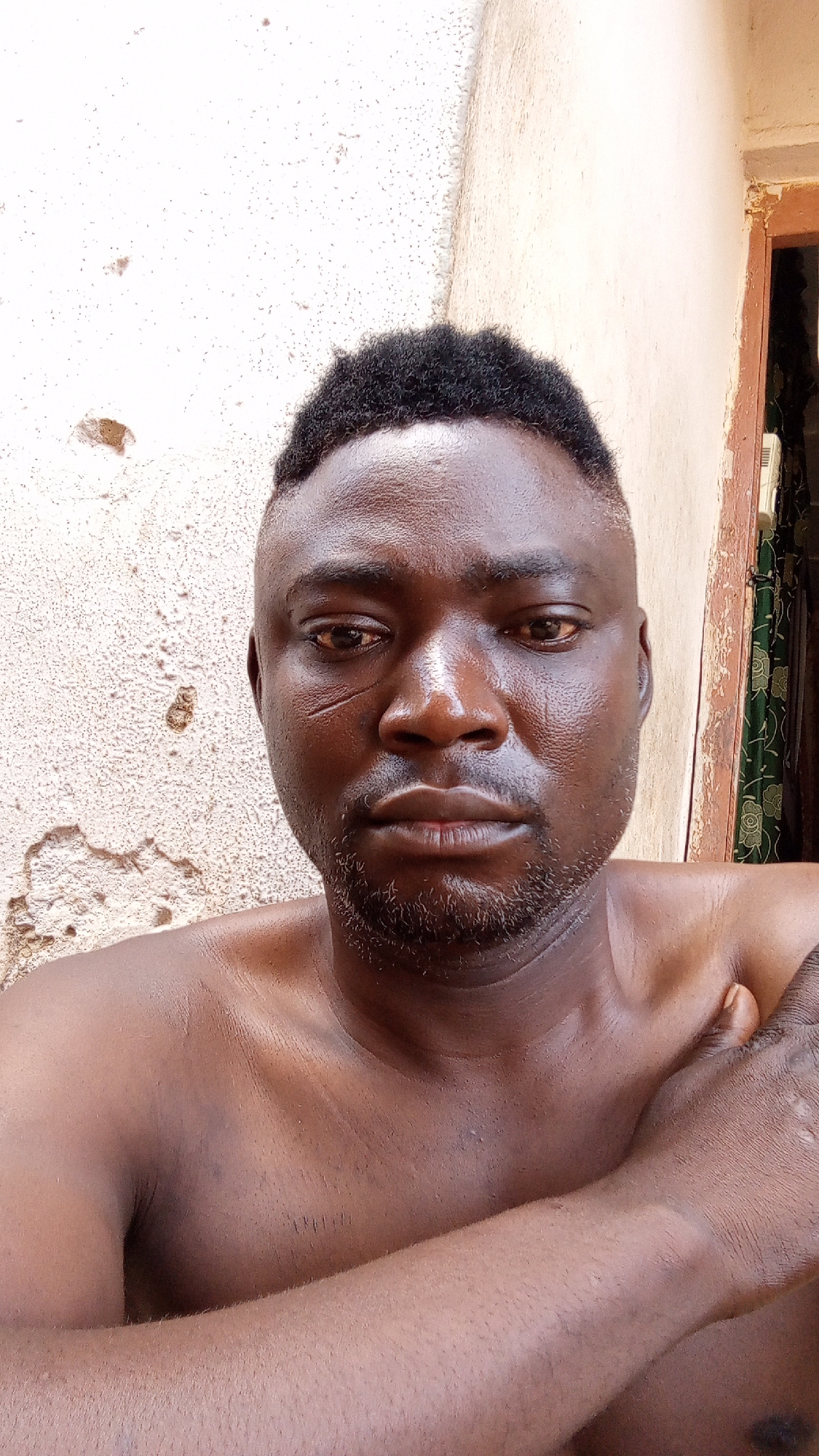 A medium close-up shot depicts a dark-skinned man with short, dark hair, a beard, and a scar on his left cheek. The man is shirtless, with his arms crossed in front of him. Behind the man is a weathered wall with damaged plaster, and a glimpse of a building with a door and curtains. The location is Sabon Tasha, Nigeria.