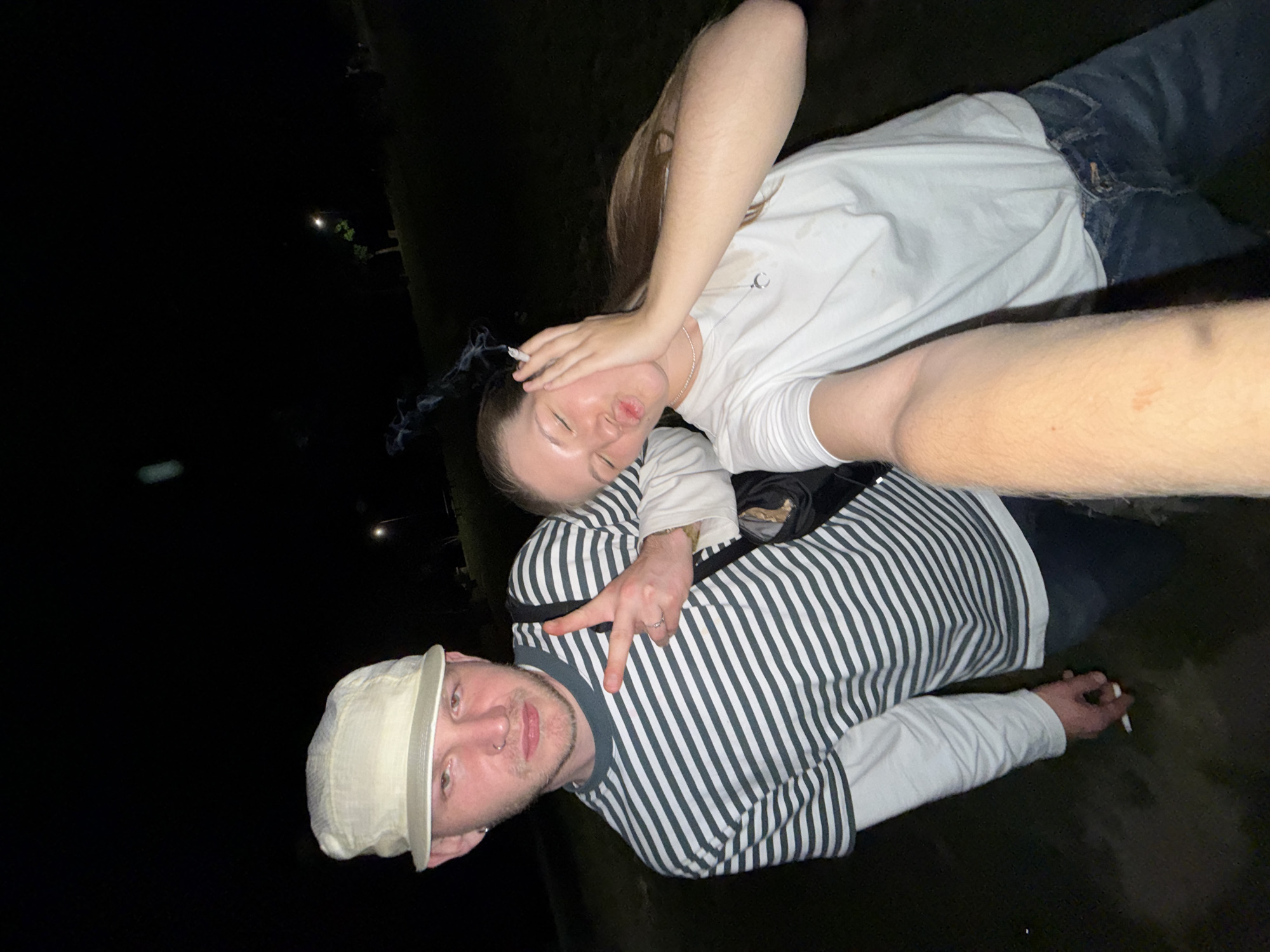 Two people are pictured outdoors at night, in what appears to be Cukarica Urban Municipality, Serbia. The lighting is dim. A person wearing a white cap and a striped shirt is lying on their back. Their hand is raised in a peace sign, and they are holding a cigarette in the other hand. A second person, wearing a white t-shirt and jeans, is lying on top of the first person, with their hand on their forehead and a cigarette in their mouth.