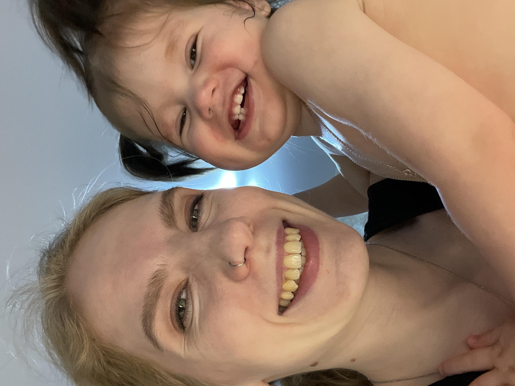 A close-up image, likely a selfie, depicts an adult female and an infant/toddler together in an indoor setting in the city of Izhevsk, Russia.
The adult female, positioned on the left side of the frame, has light blonde or light brown hair, green-blue eyes, and a silver nose ring on her left nostril. She exhibits a wide smile, exposing upper and lower teeth with a yellowed appearance. She wears a dark-colored garment, possibly a t-shirt, and a thin silver chain necklace. Her right arm is partially visible, supporting the child.
The infant/toddler, positioned on the right, has dark brown hair styled into two small pigtails. The child has brown eyes and is smiling widely, revealing developing teeth and a visible reddish mark on the left side of the forehead. The child appears to be shirtless or minimally clothed, exposing the upper chest and shoulder, and also wears a thin silver chain necklace.
Both individuals are facing and looking directly into the camera. The background is uniformly light-colored, out of focus, and lacks distinct features, suggesting an interior space with ambient lighting.